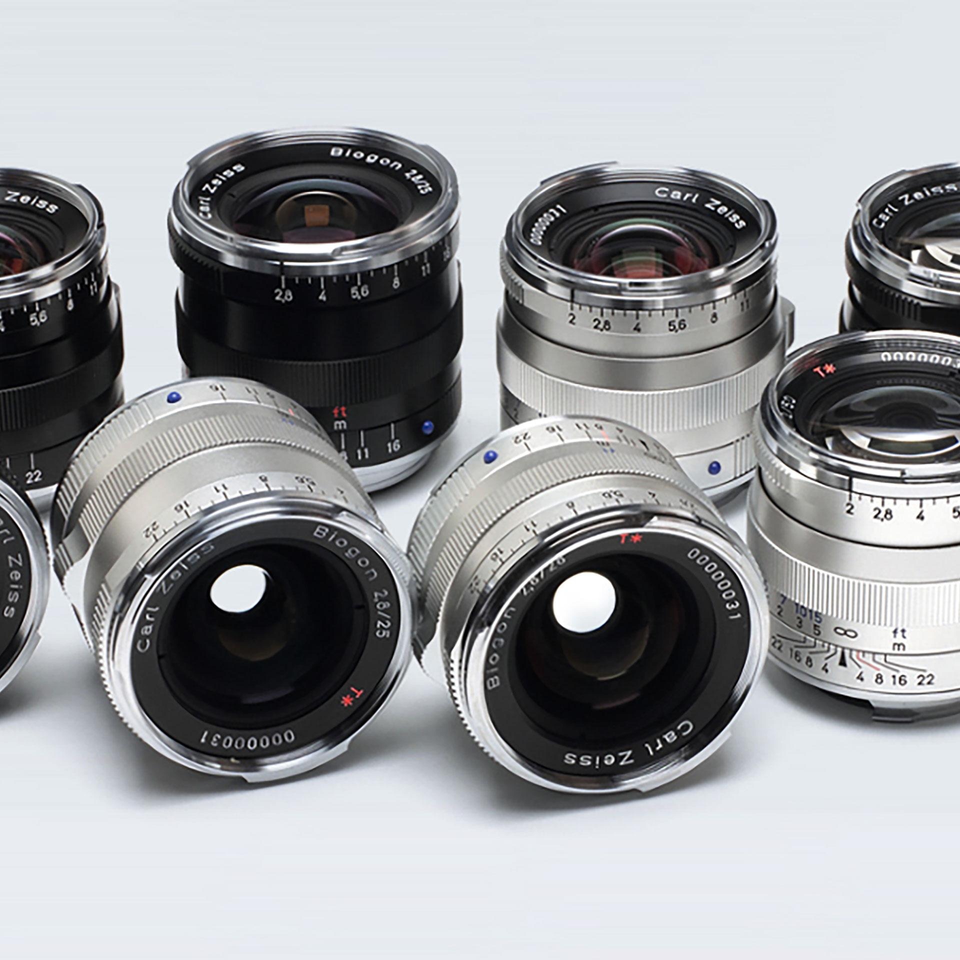 A collection of nine ZEISS camera lenses in black and silver finishes arranged on a light gray background.