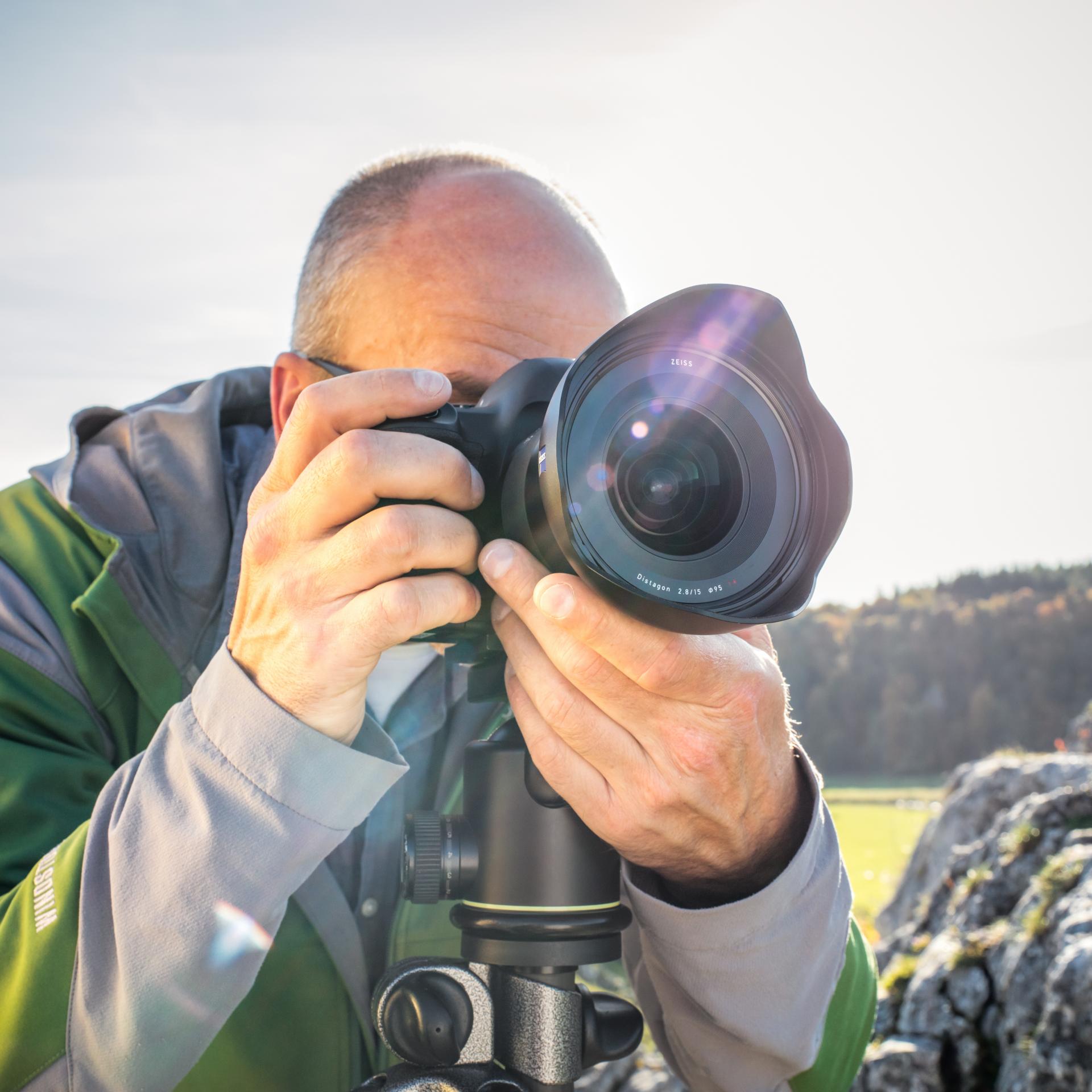 A photographer is taking a picture outdoors with a camera equipped with a ZEISS wide-angle lens mounted on a tripod.