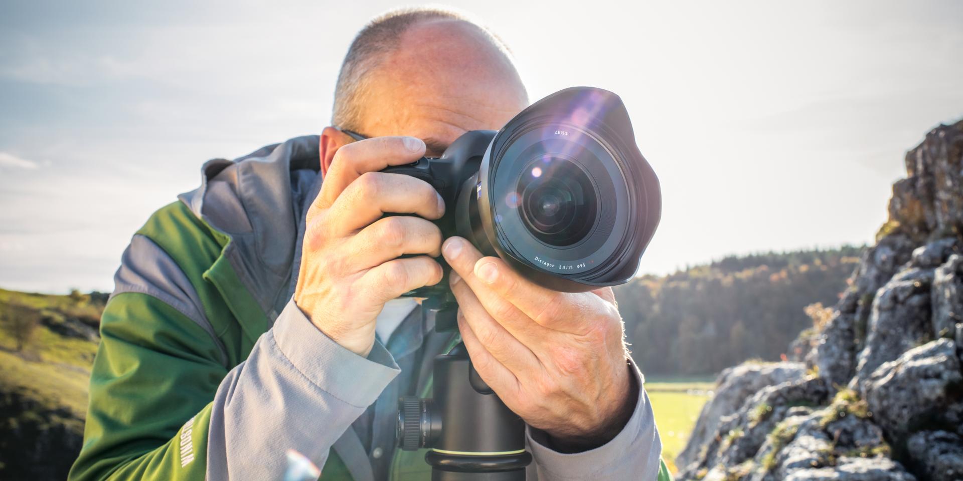 A photographer is taking a picture outdoors with a camera equipped with a ZEISS wide-angle lens mounted on a tripod.