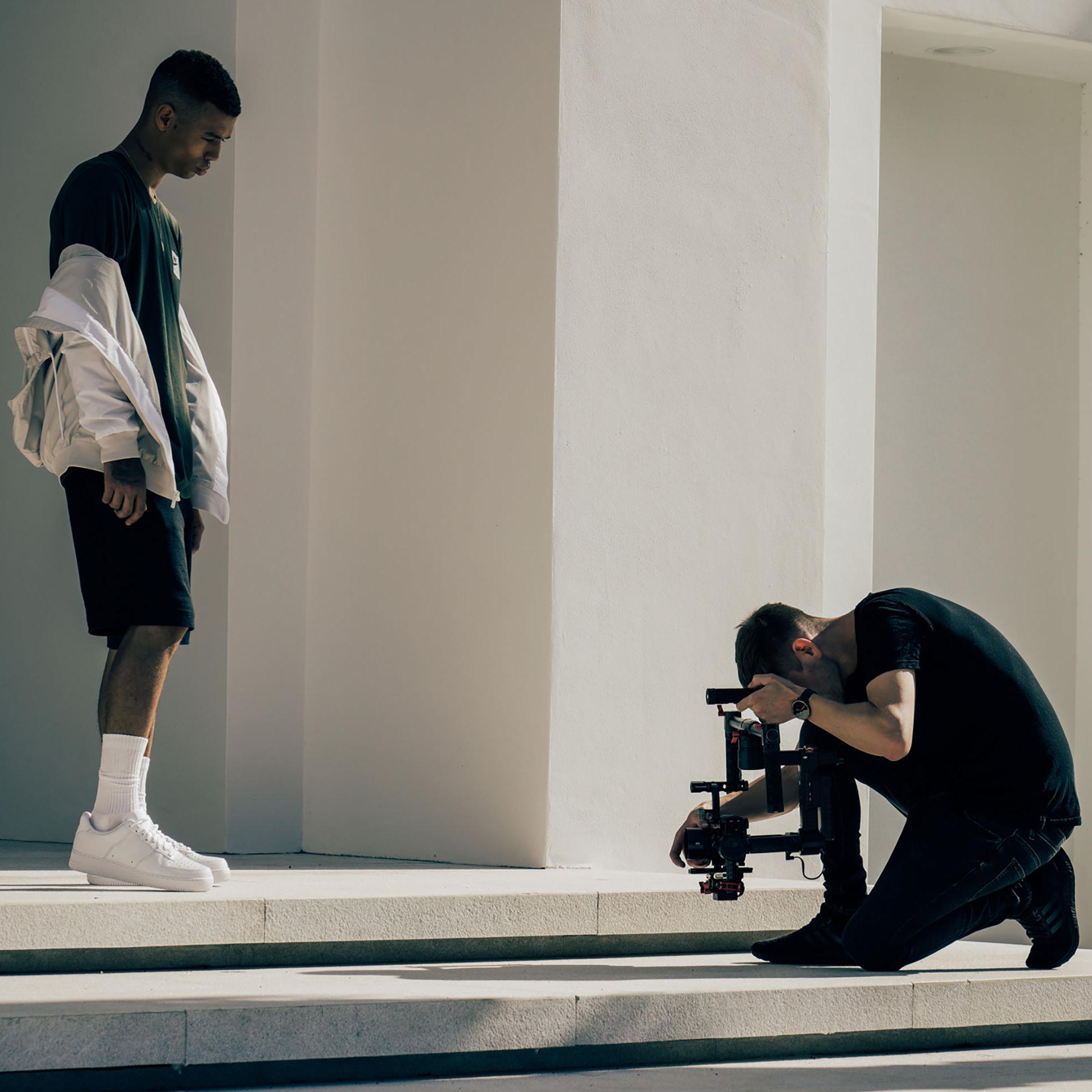 A videographer kneels while filming a man standing on white steps outside a modern building, using a professional camera setup with ZEISS lenses.