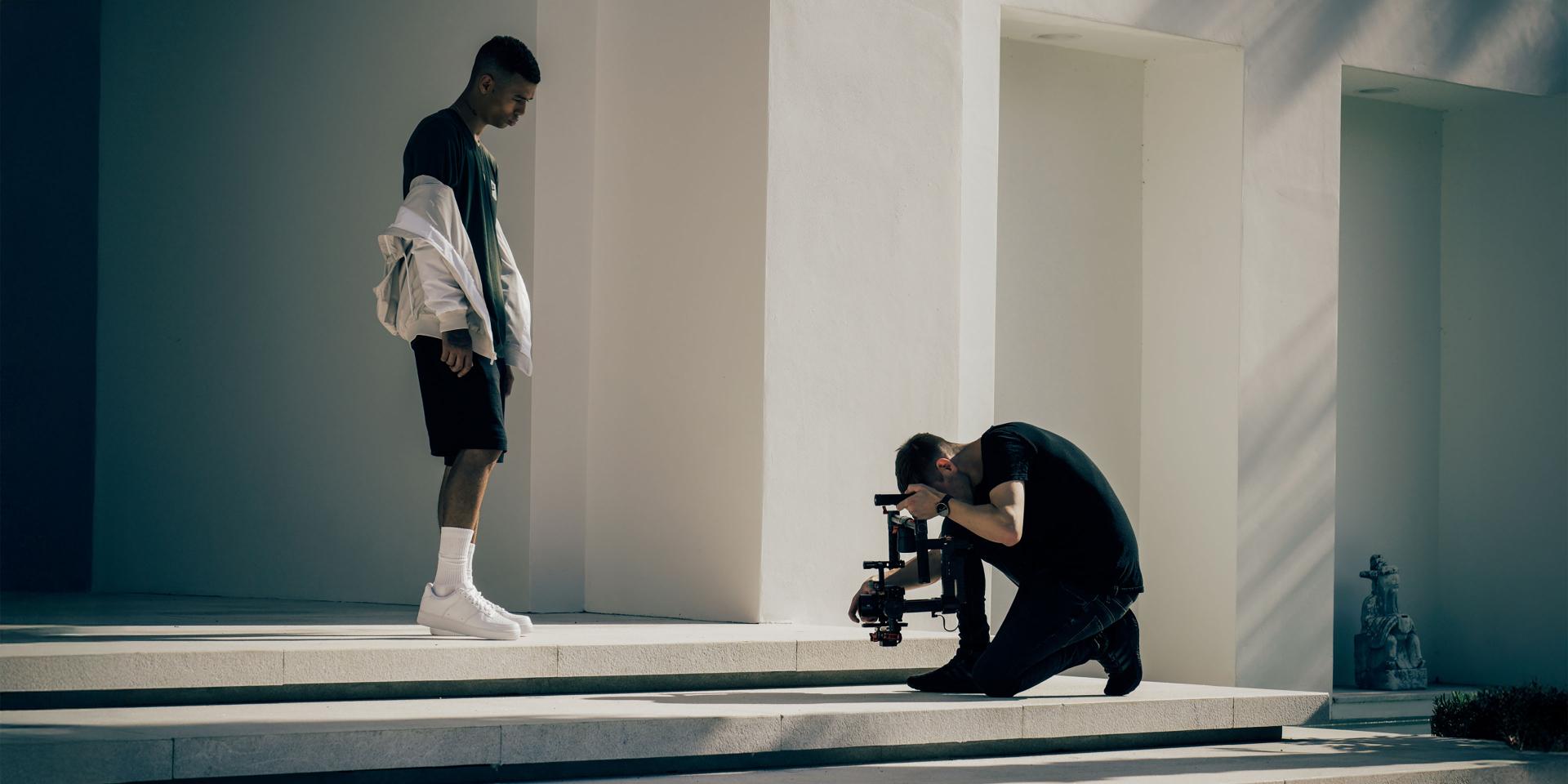 A videographer kneels while filming a man standing on white steps outside a modern building, using a professional camera setup with ZEISS lenses.