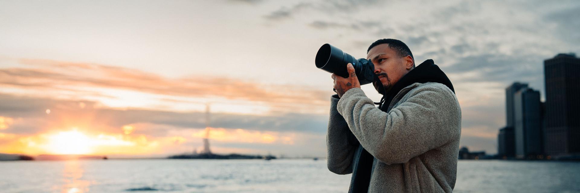 Man taking a photo with a ZEISS camera lens at sunset by the waterfront, with city buildings in the background.