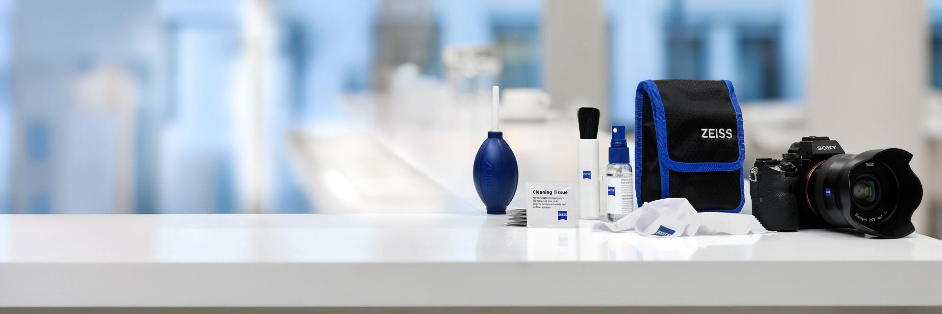 A ZEISS camera cleaning kit with various cleaning tools, a carrying pouch, and a camera is displayed on a white surface with a blurred background.