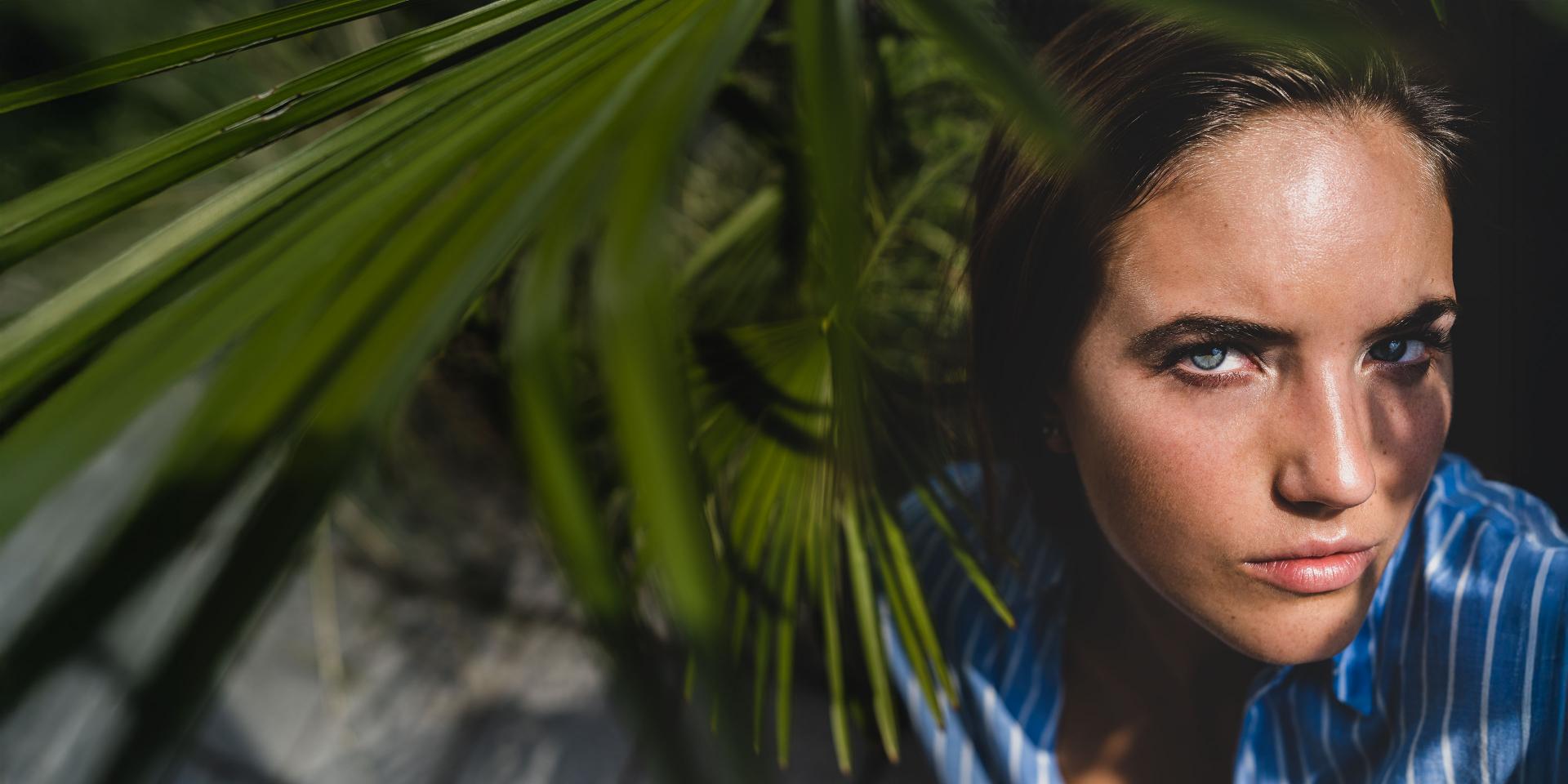 A woman in a blue striped shirt looks intently at the camera through palm leaves casting shadows on her face.