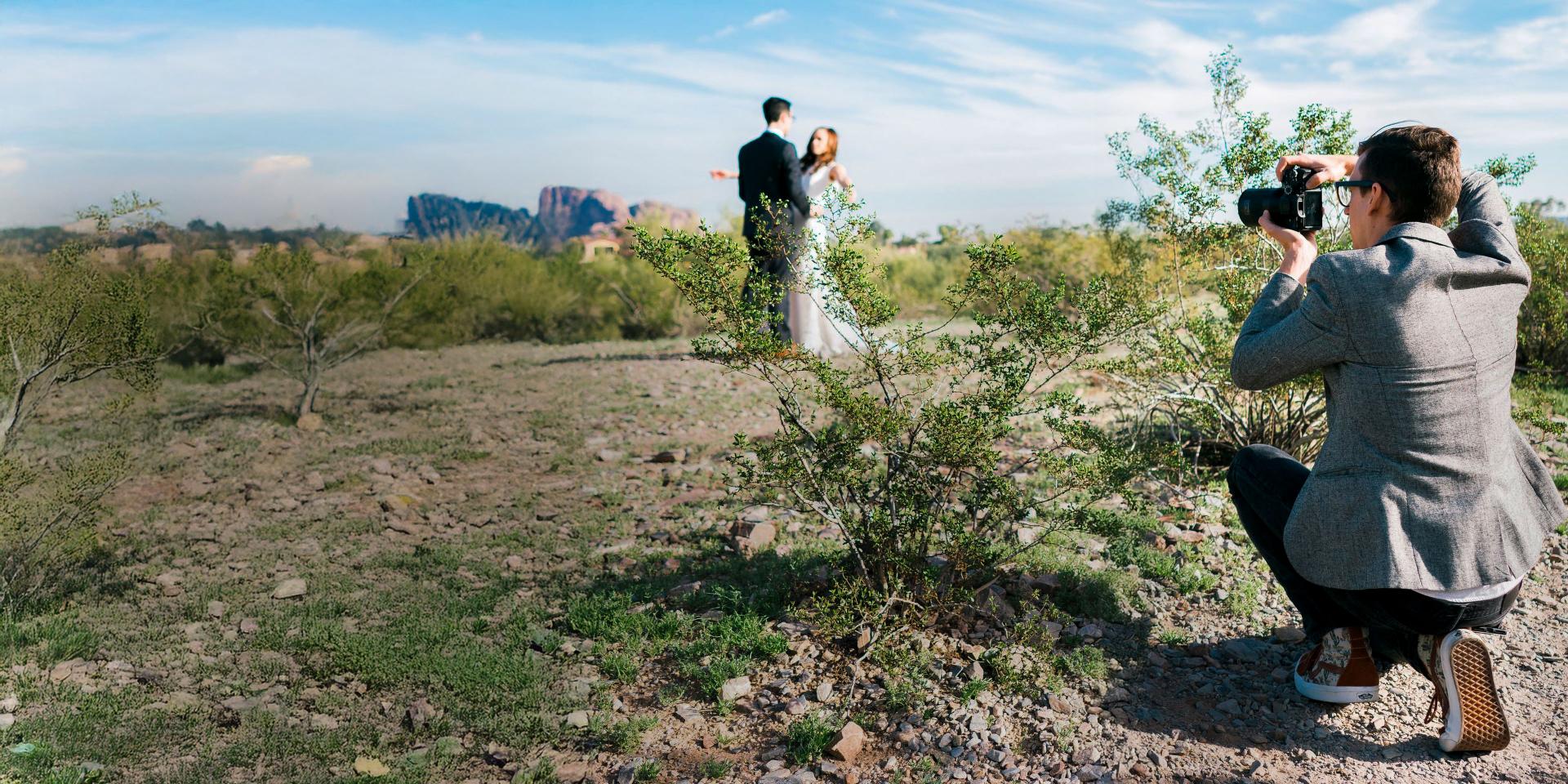 A photographer using a ZEISS camera lens captures a wedding couple posing in a scenic desert landscape.