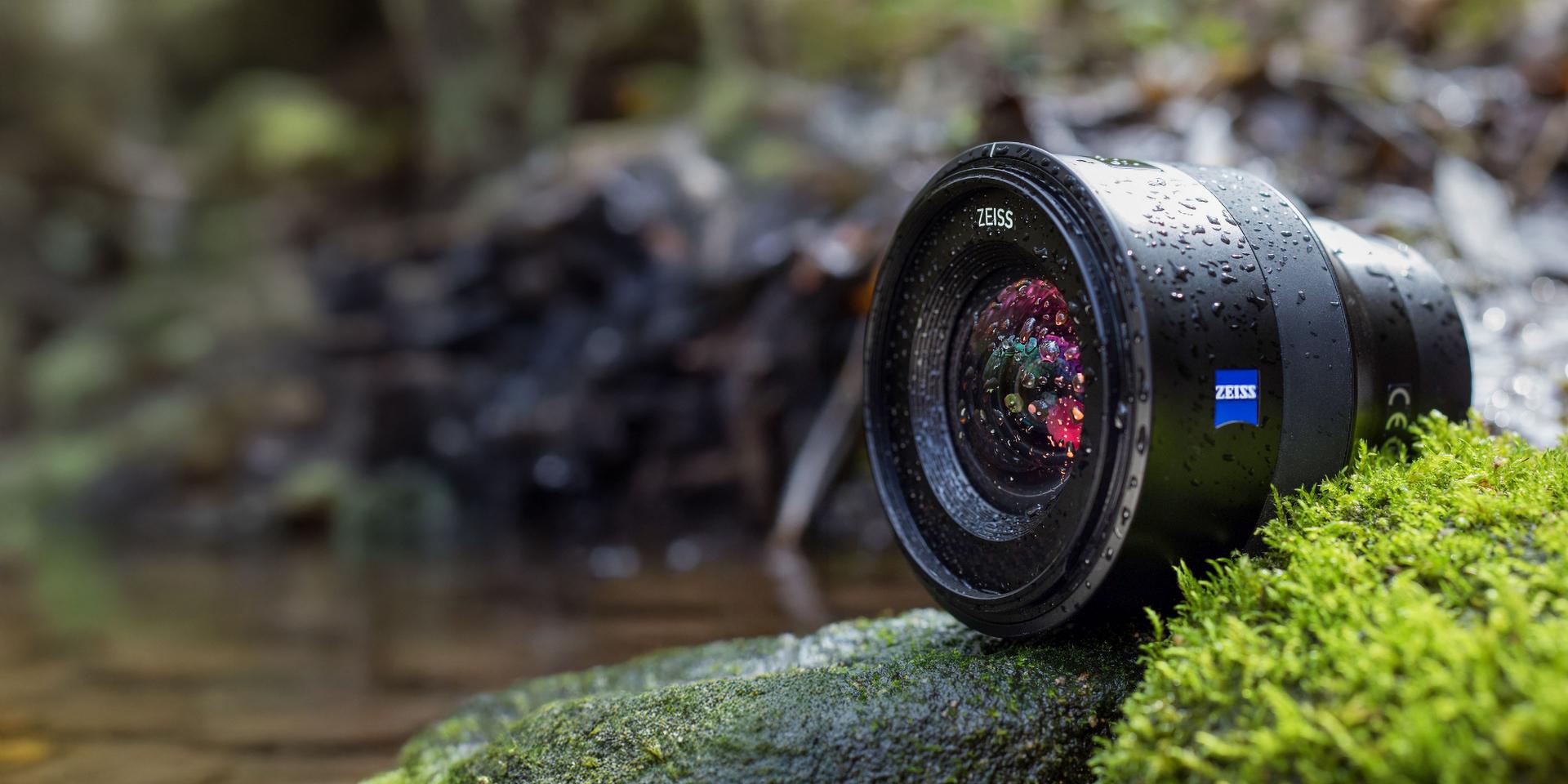 A ZEISS camera lens covered in water droplets rests on a mossy rock beside a stream in a natural outdoor setting.