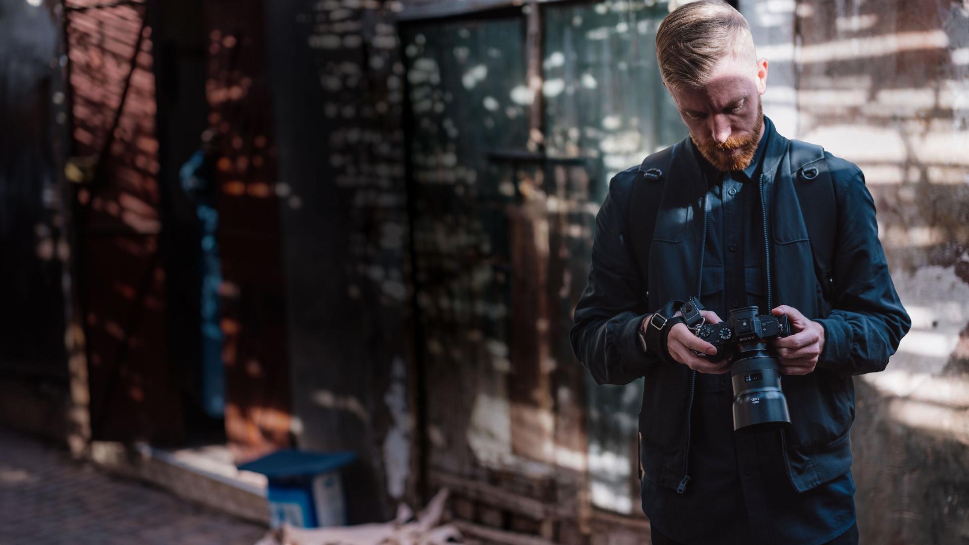 A photographer in a dark jacket is examining a camera equipped with a ZEISS lens in an urban outdoor setting.