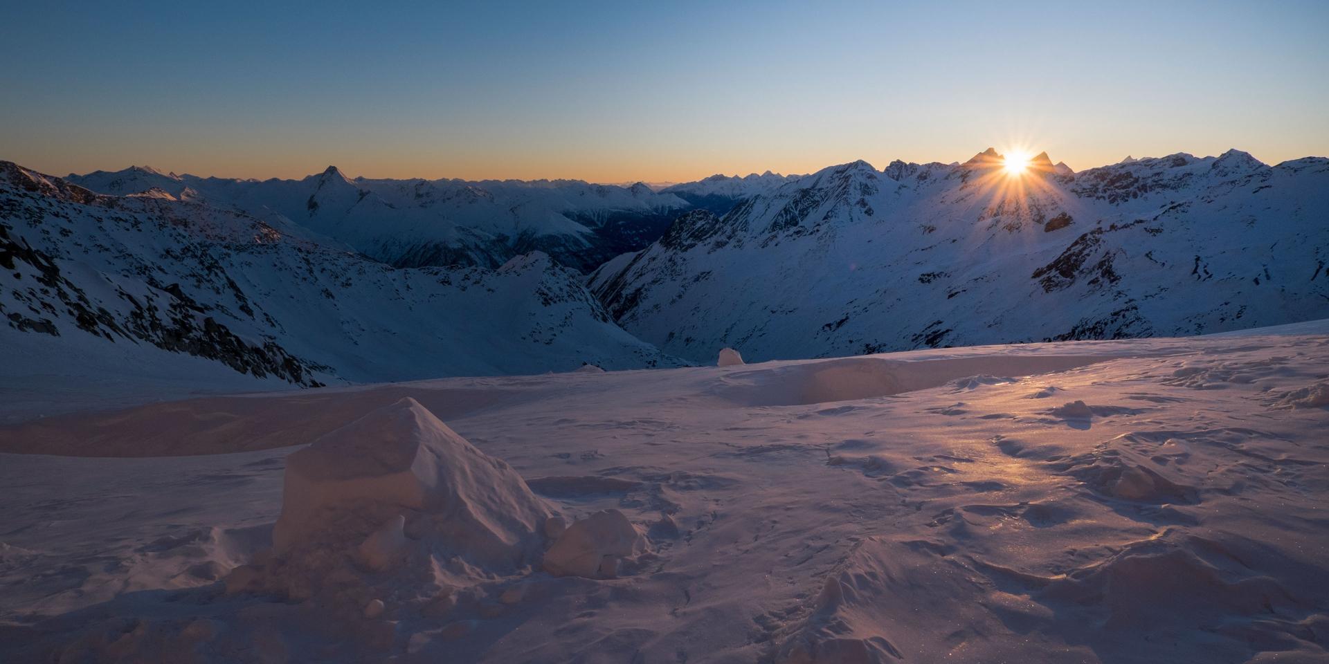 Snow-covered mountain slope with rippled texture illuminated by the setting sun behind distant peaks.