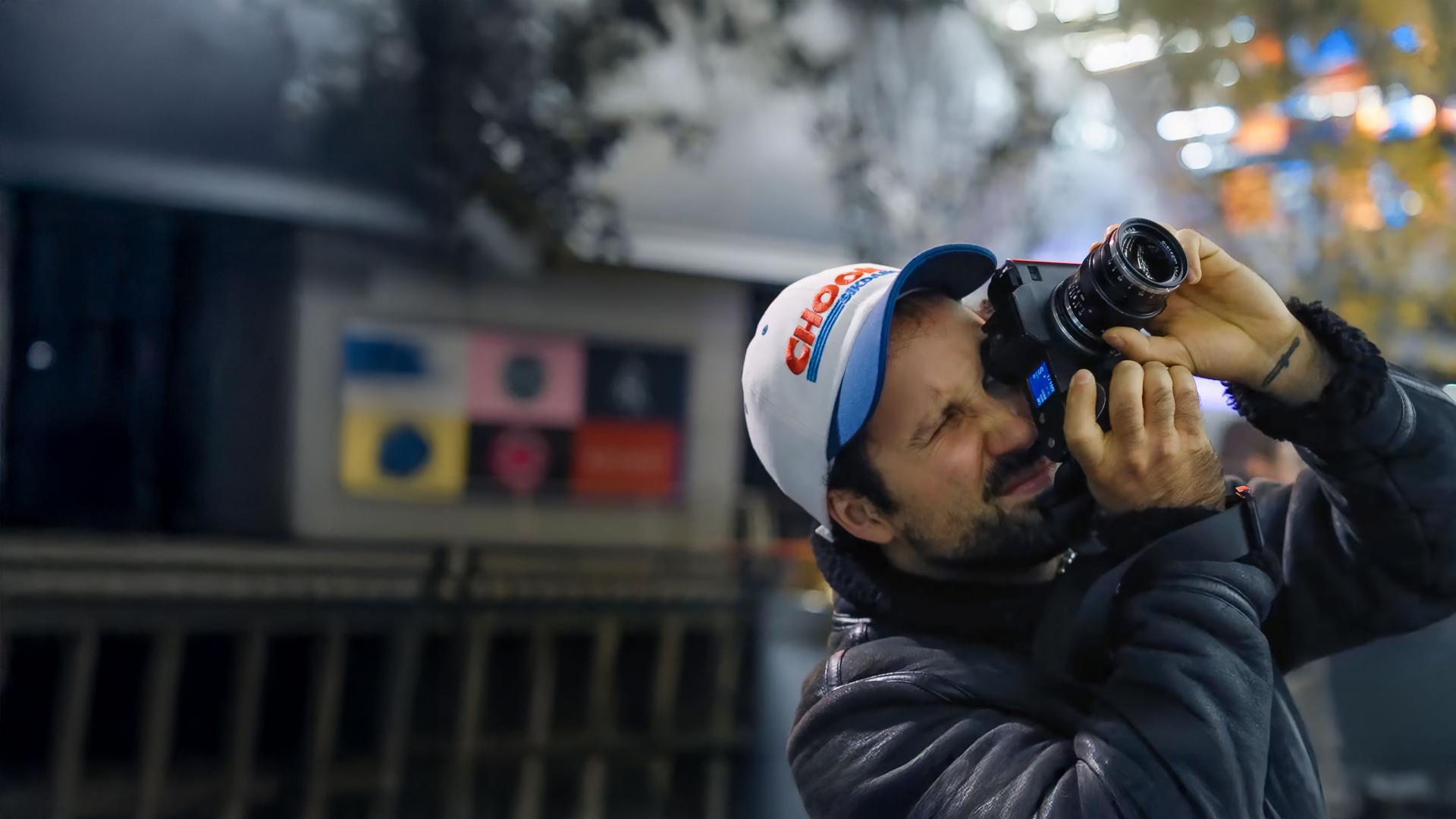 A man wearing a cap and jacket is using a camera equipped with a ZEISS lens in an indoor setting.