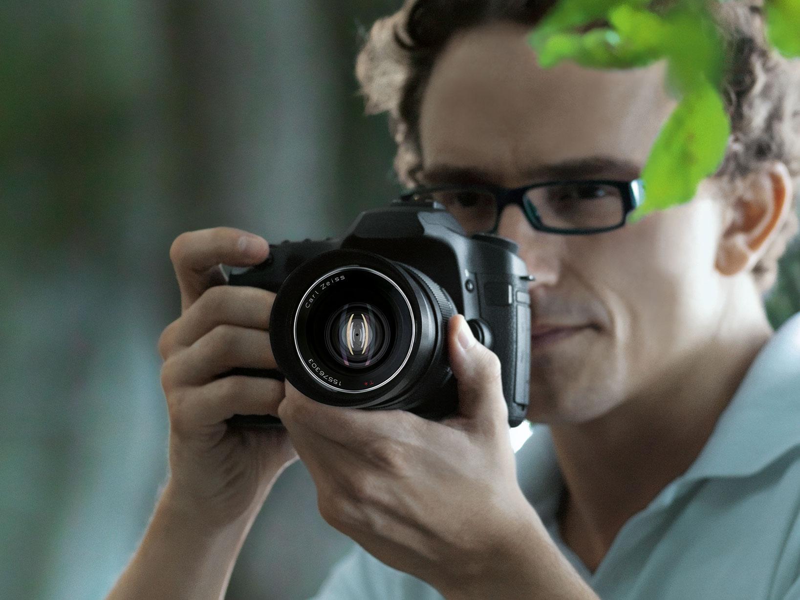Man wearing glasses holding a DSLR camera equipped with a ZEISS lens, focusing on a shot outdoors.