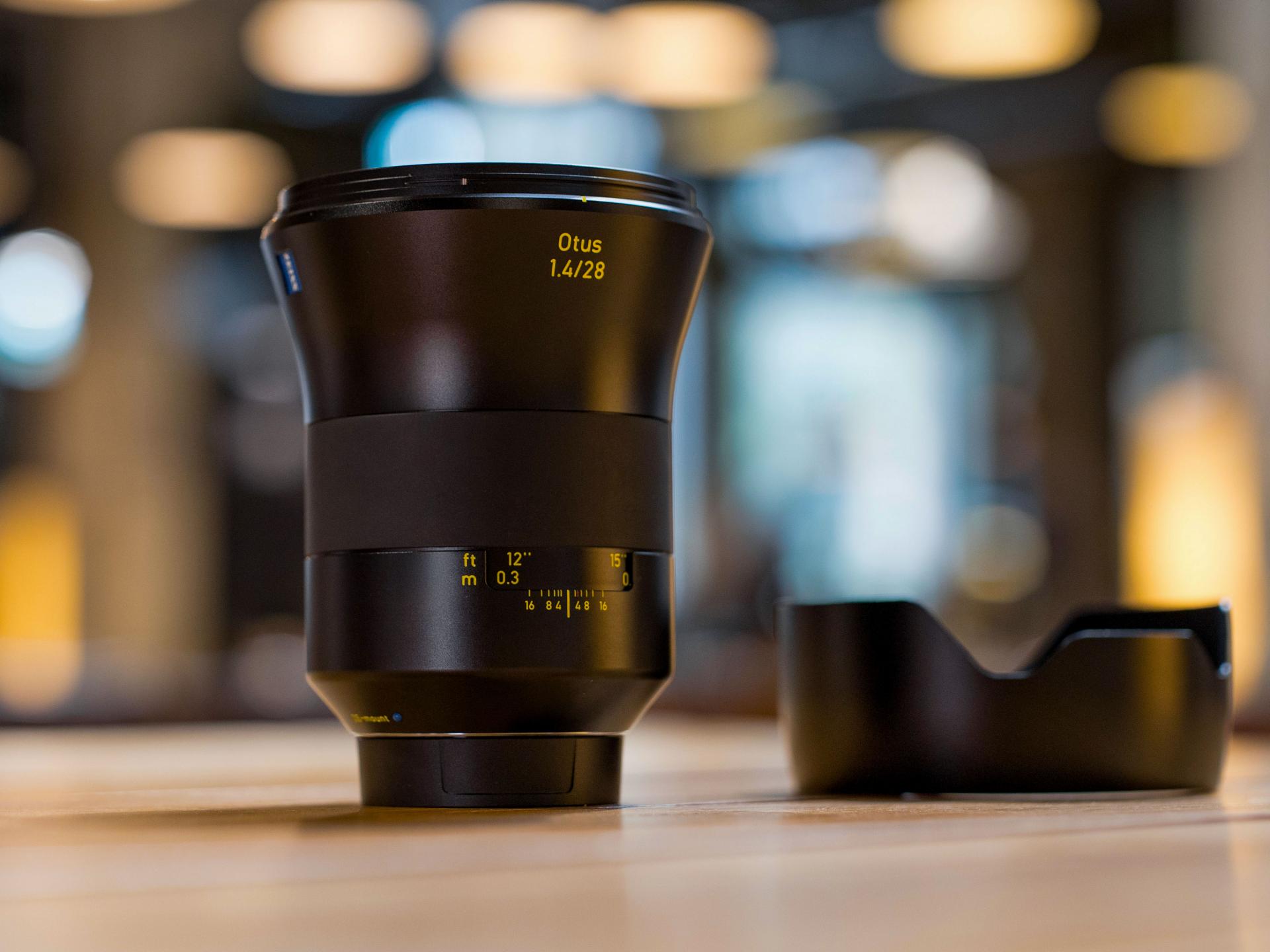 ZEISS Otus 1.4/28 camera lens standing upright on a wooden surface with its lens hood placed beside it, set against a softly blurred indoor background.
