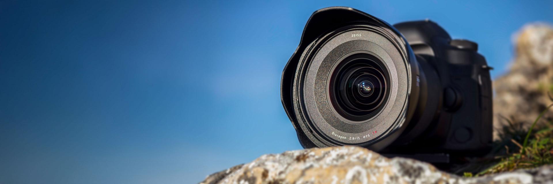 Close-up of a professional camera with a ZEISS lens resting on a rock against a clear blue sky.