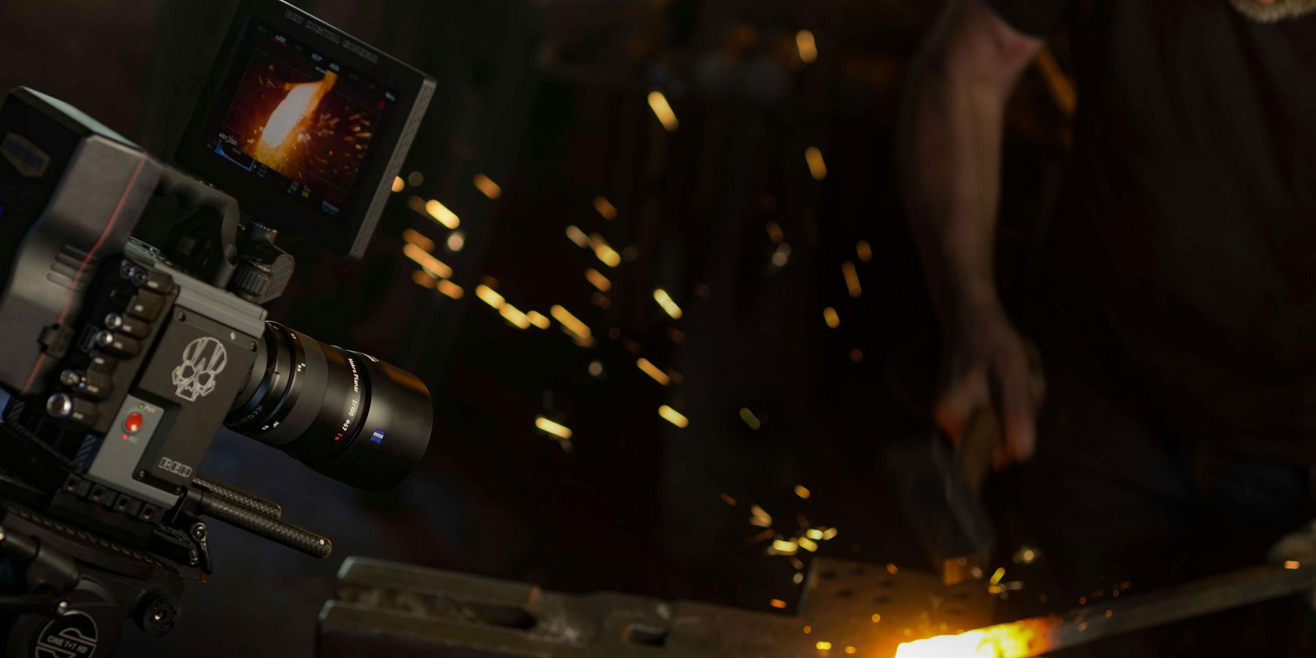A filmmaker records a blacksmith at work near a furnace using a professional camera equipped with a ZEISS lens.