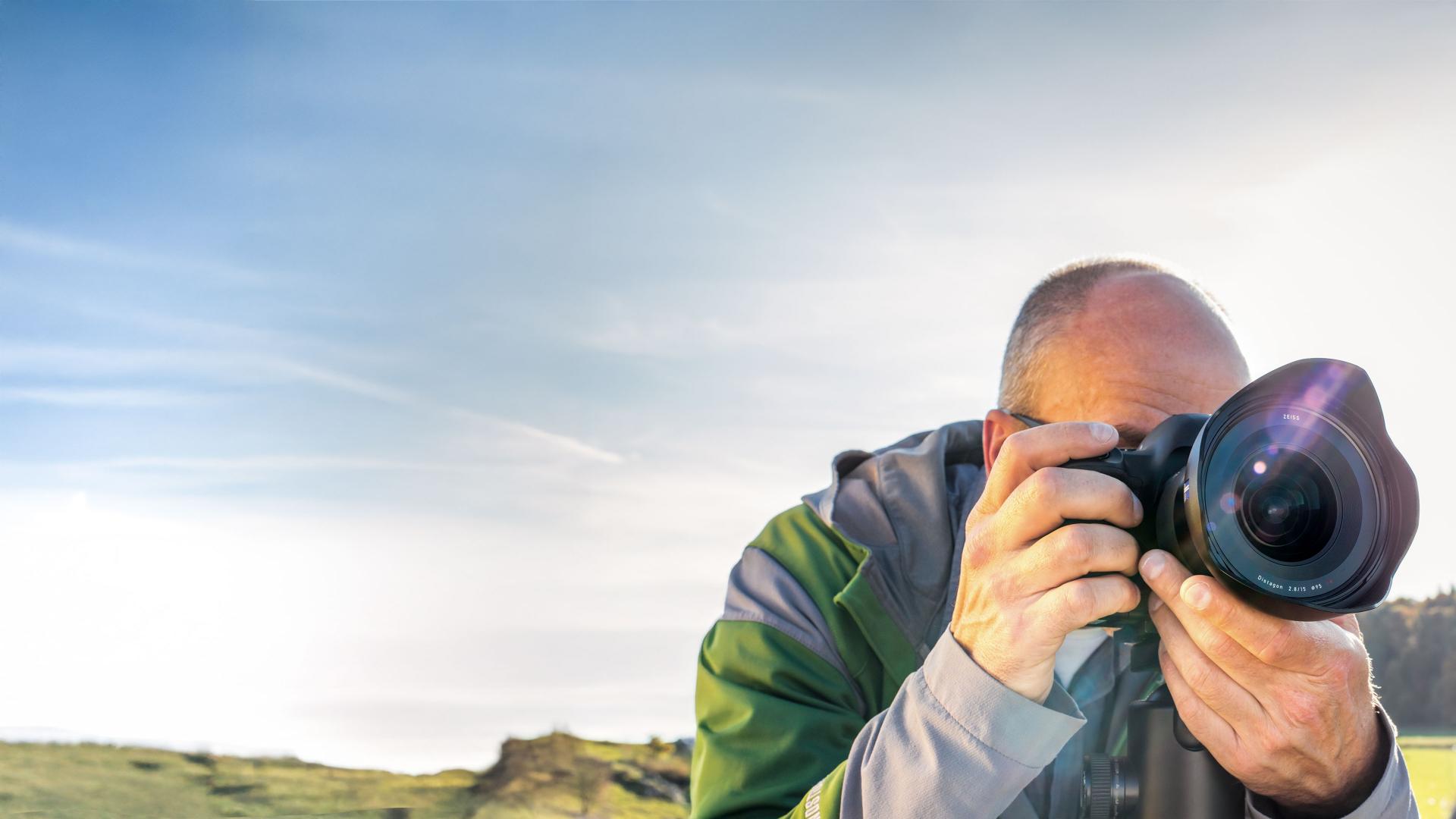 A man in outdoor clothing is taking a photo with a professional camera equipped with a ZEISS lens in a natural landscape setting.