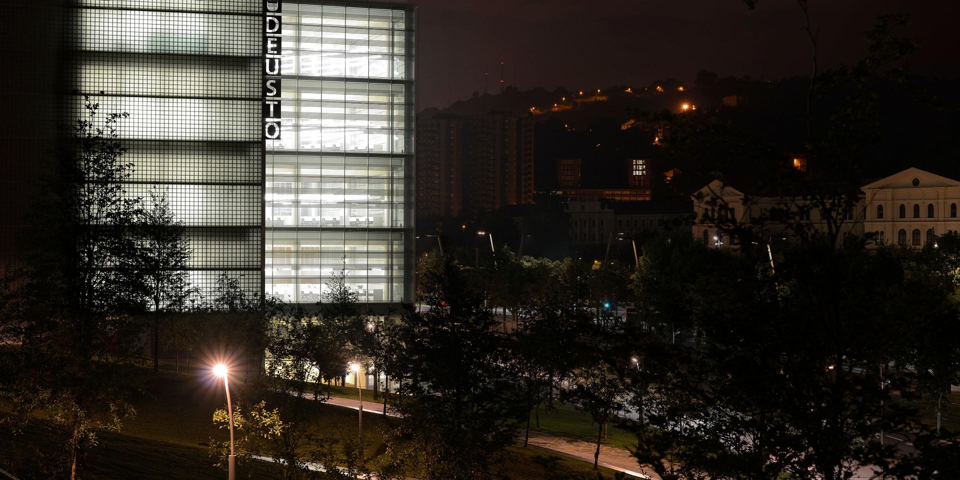 Modern glass-fronted building illuminated at night, surrounded by trees and city lights.