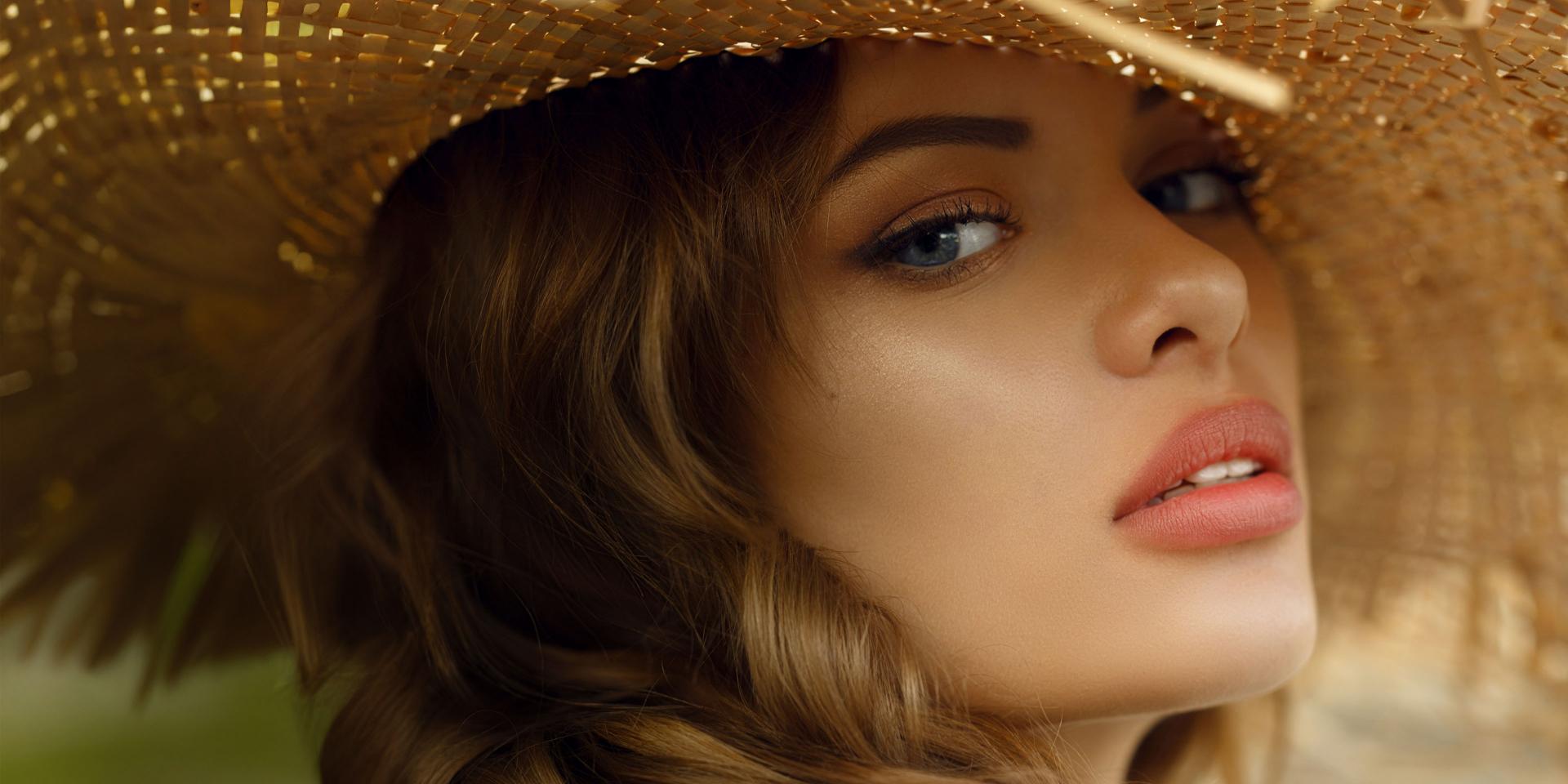 A close-up portrait captures a young woman with long, wavy brown hair, partially obscured by a large, wide-brimmed straw hat, her piercing blue eyes and subtly made-up face creating a striking contrast against the warm tones of the hat and her skin.