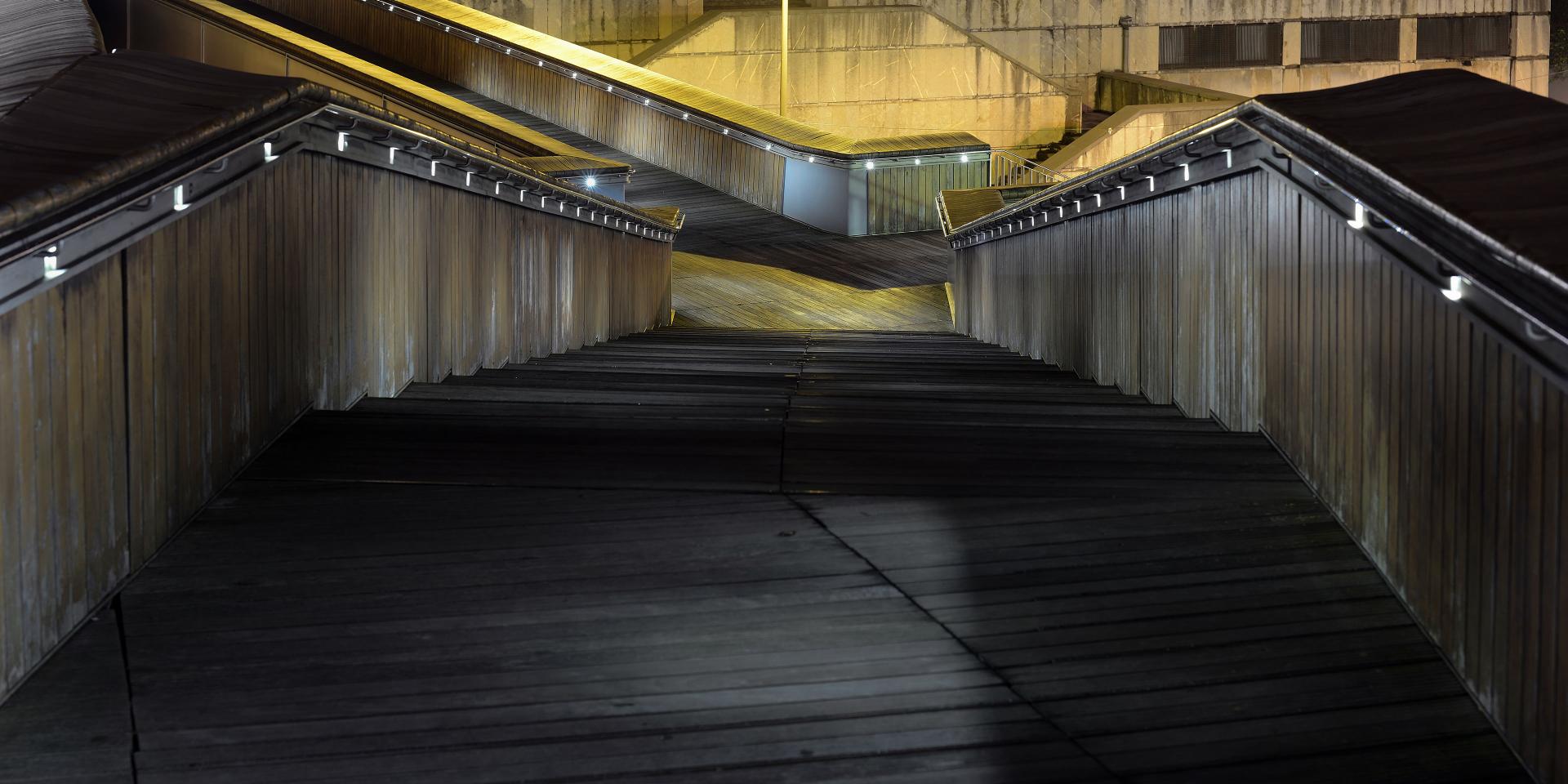 Nighttime view of a modern, multi-level wooden pedestrian ramp with metal railings, illuminated by streetlights.