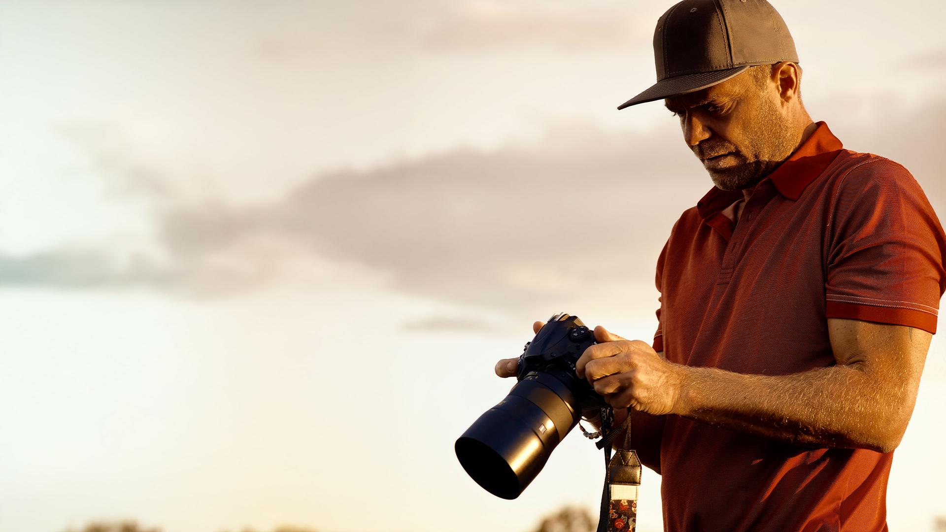 A man wearing a cap examines a camera equipped with a ZEISS lens outdoors during golden hour.