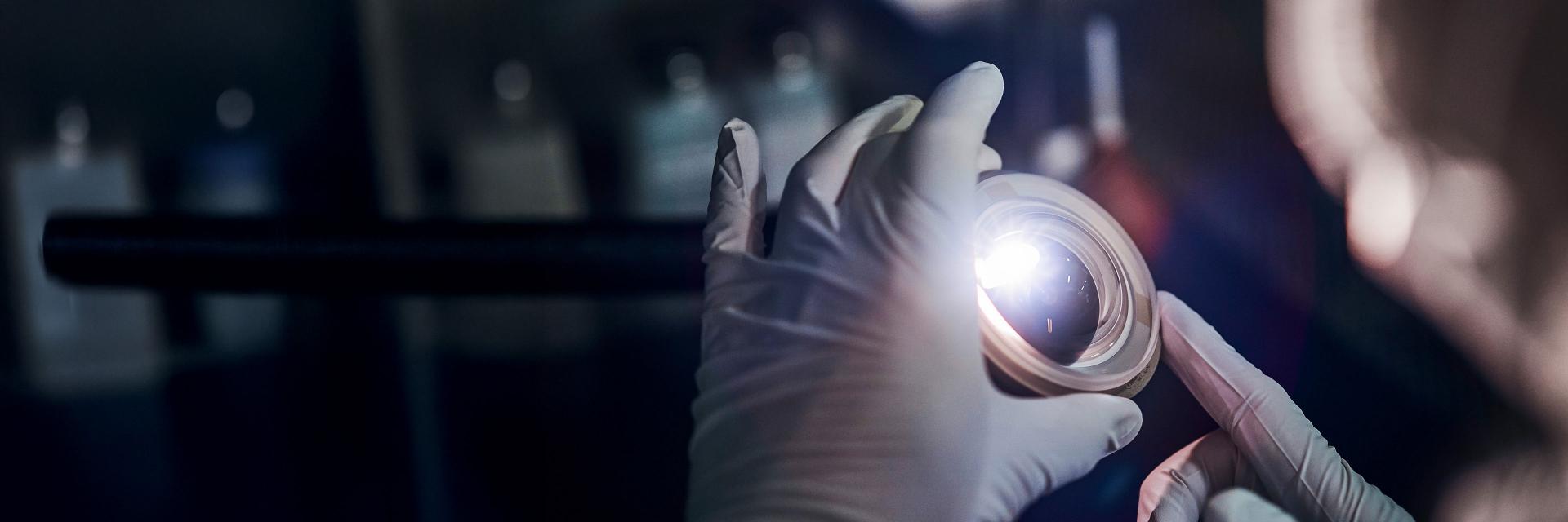 Gloved hands inspecting a ZEISS optical lens under focused light in a laboratory setting.