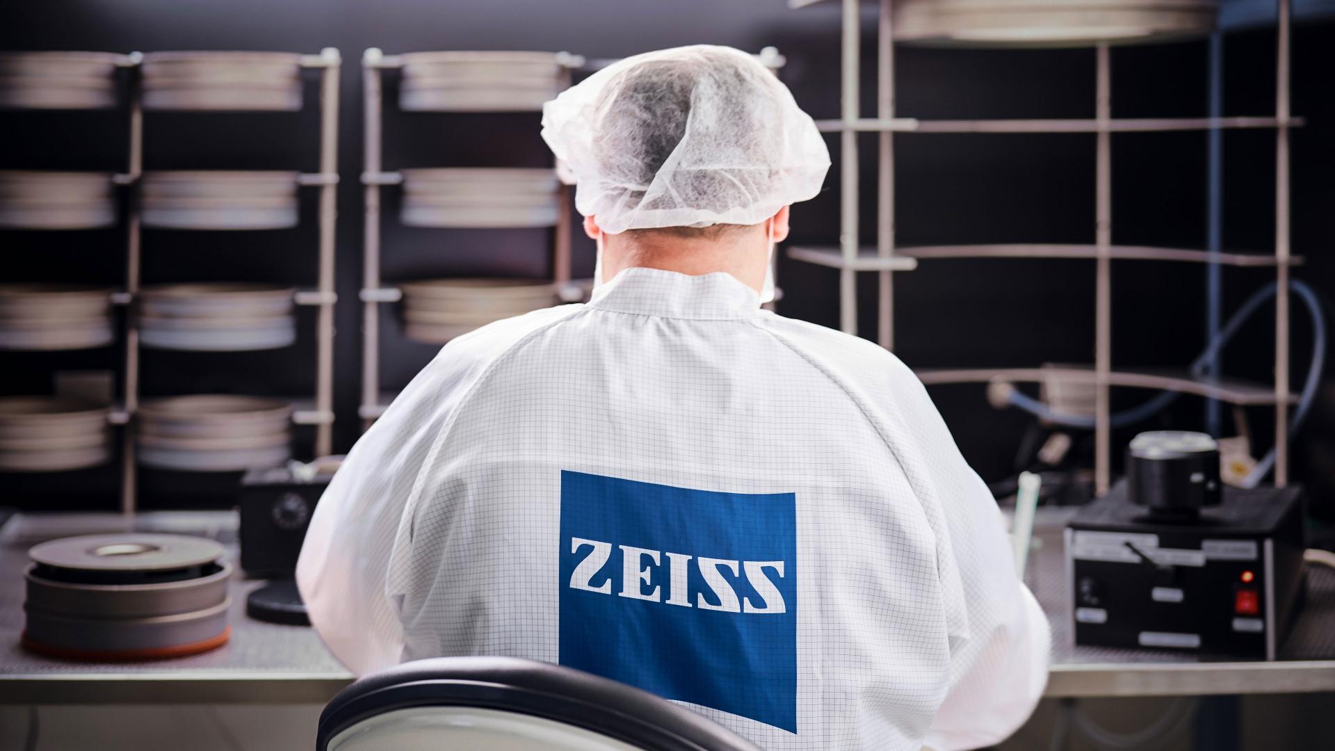 Technician in a cleanroom environment wearing a ZEISS lab coat and hairnet, working with optical equipment and materials.