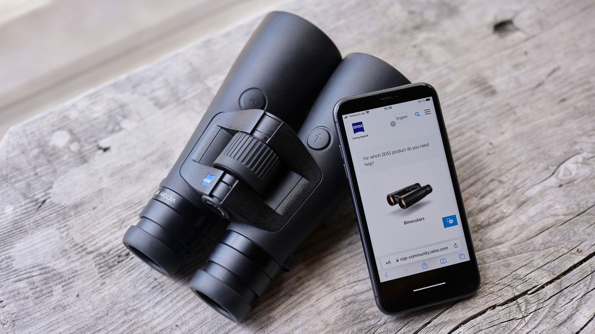 A pair of black ZEISS binoculars placed next to a smartphone displaying a ZEISS product page on a wooden surface.