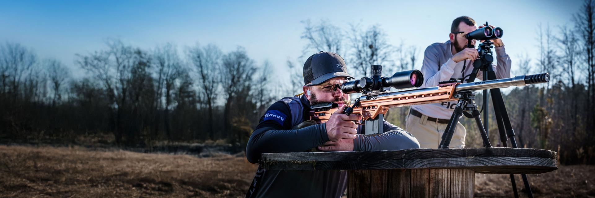 Two men are outdoors using ZEISS optics, one aiming with a rifle equipped with a ZEISS scope and the other observing through ZEISS binoculars on a tripod.