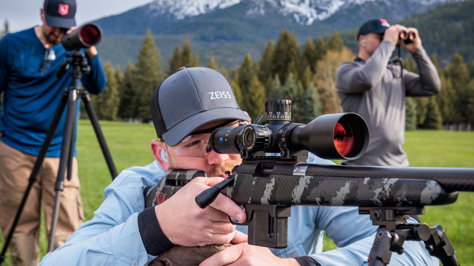 Three men are outdoors using ZEISS optics equipment, with one aiming a rifle scope, another using a spotting scope on a tripod, and the third looking through binoculars against a backdrop of trees and mountains.