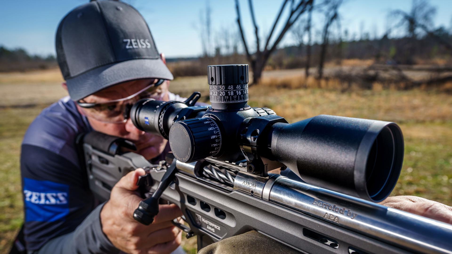 A person aiming a precision rifle equipped with a ZEISS riflescope in an outdoor shooting range.