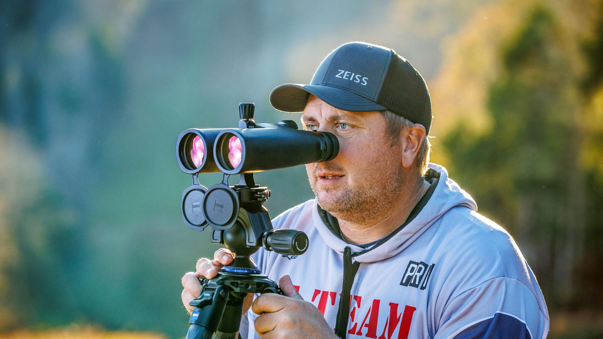 A man wearing a ZEISS cap is using a ZEISS binocular mounted on a tripod in an outdoor setting.