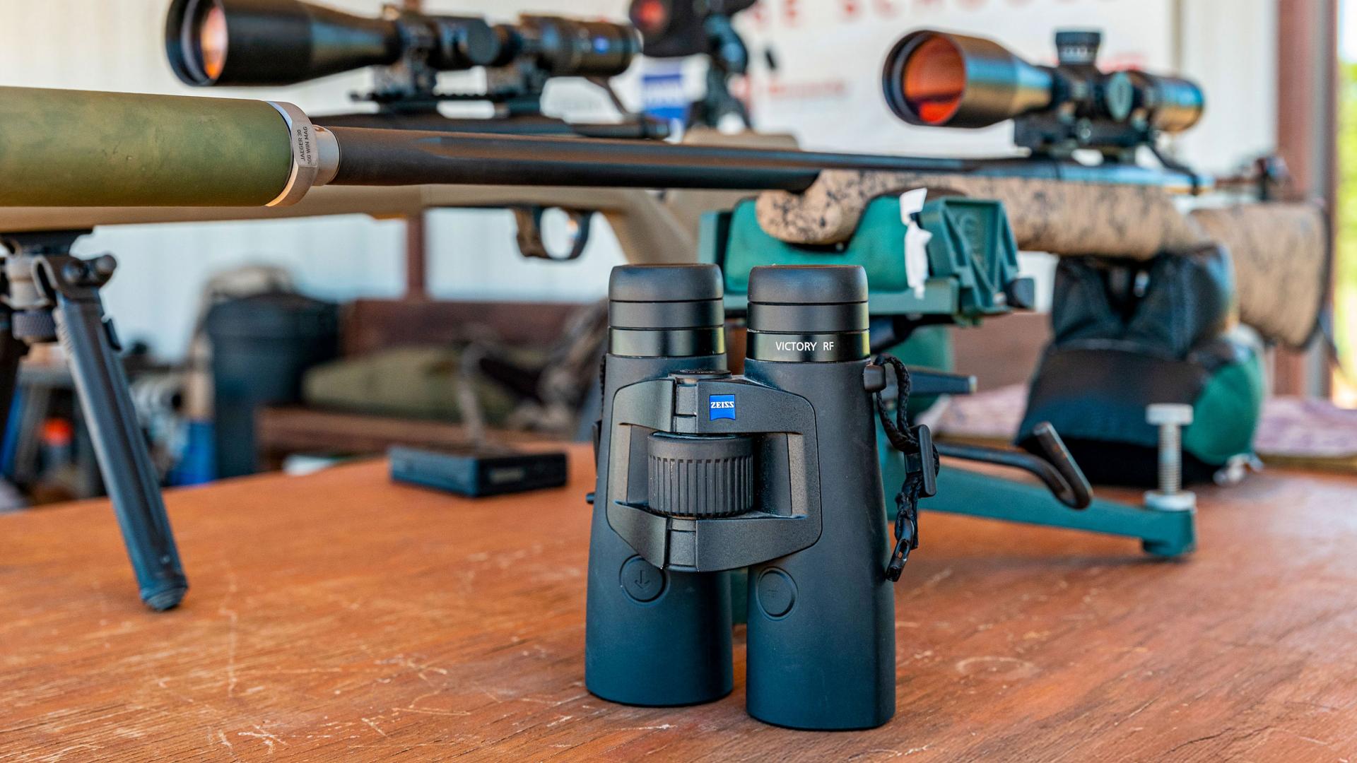 A pair of ZEISS binoculars is placed on a wooden table in front of precision rifles mounted on tripods at an outdoor shooting range.
