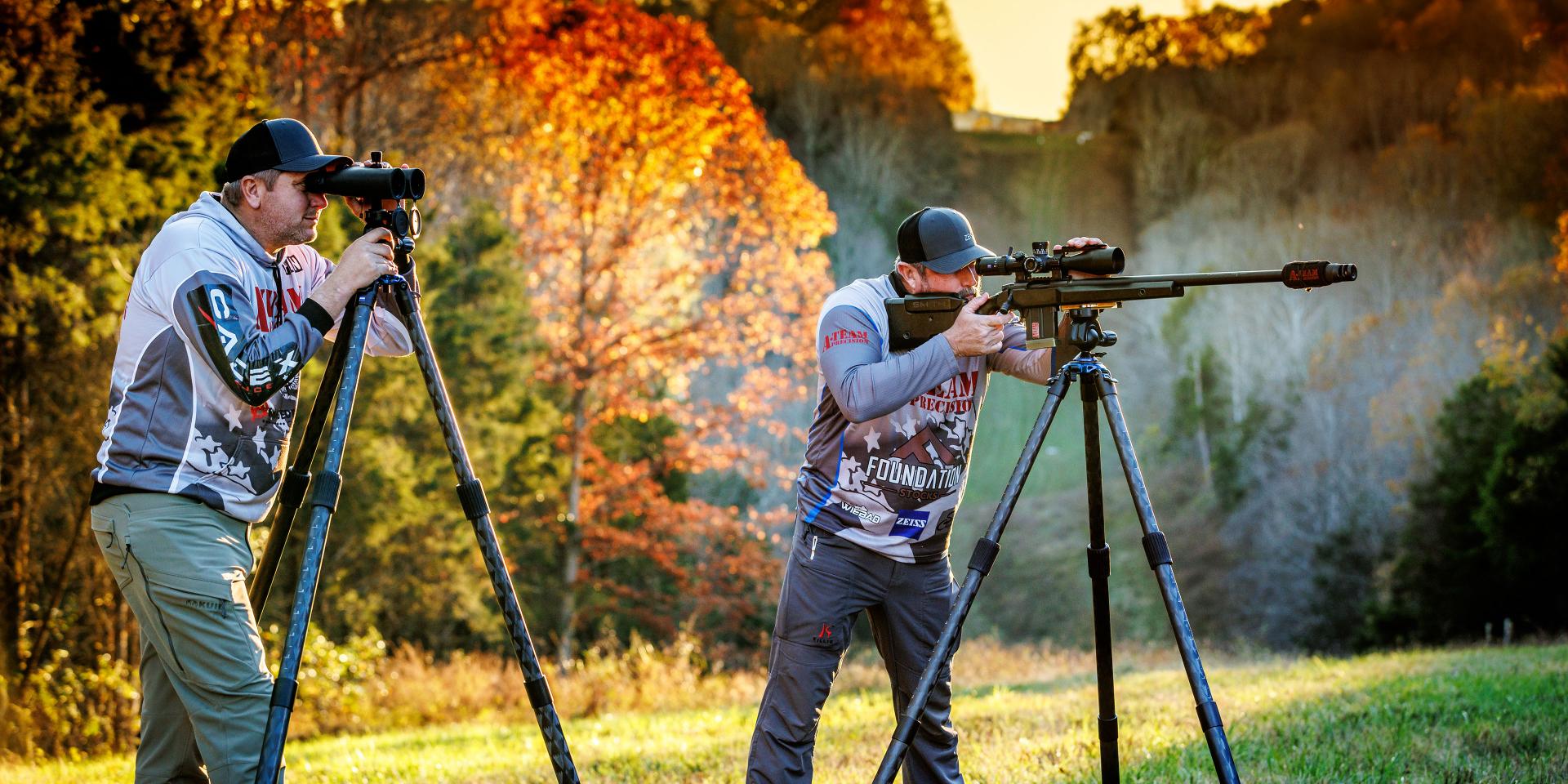 Two men using ZEISS optics mounted on tripods for spotting and precision shooting in a scenic outdoor setting with autumn foliage.