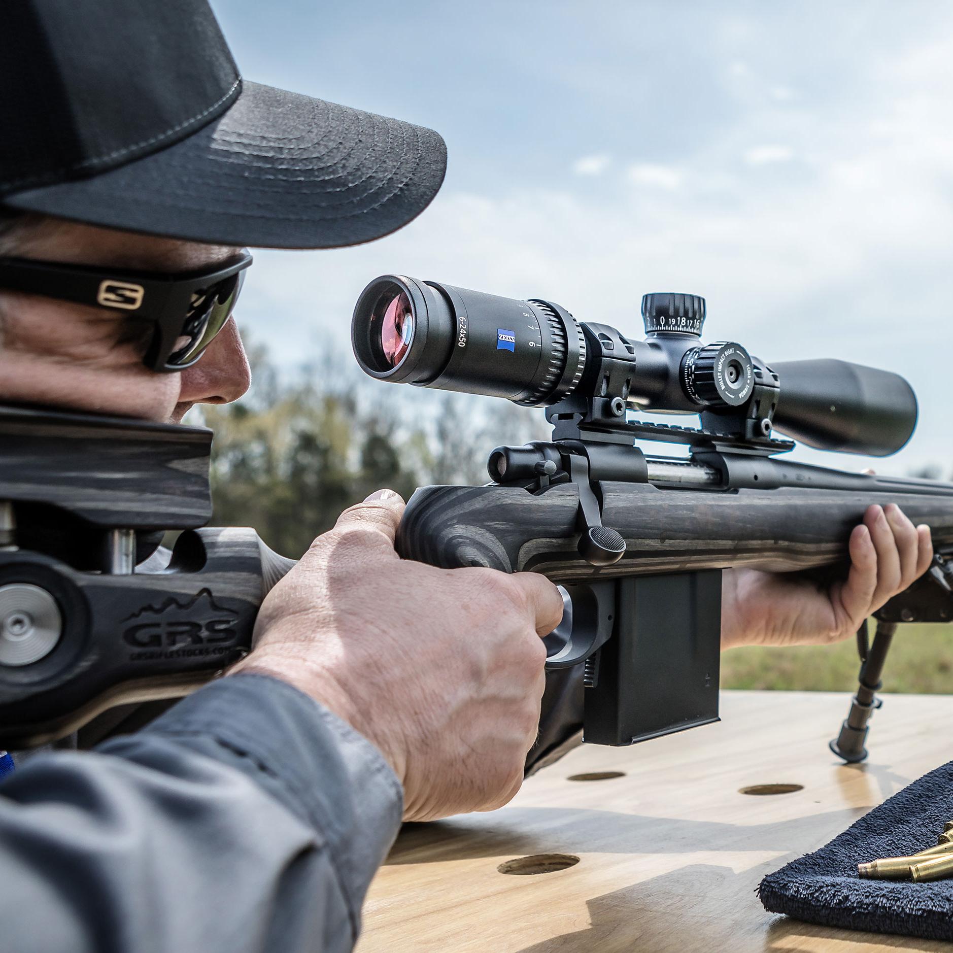A person aiming a rifle equipped with a ZEISS riflescope at an outdoor shooting range, with ZEISS binoculars and ammunition placed on a wooden table.