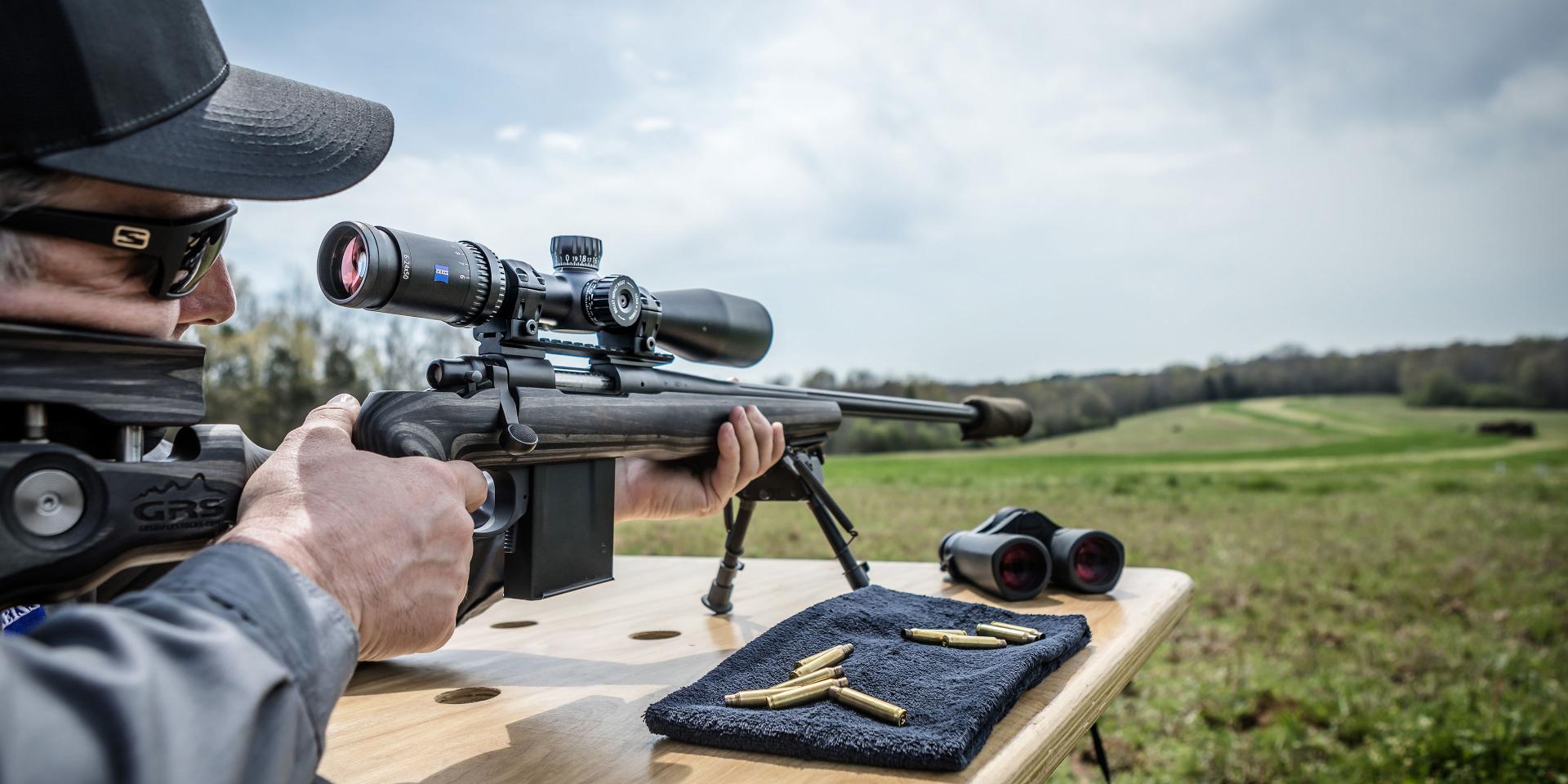 A person aiming a rifle equipped with a ZEISS riflescope at an outdoor shooting range, with ZEISS binoculars and ammunition placed on a wooden table.