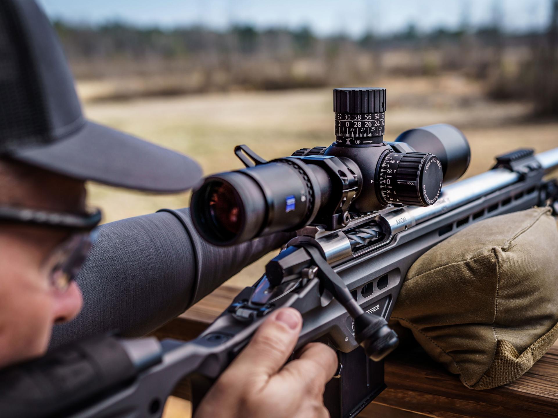 A person aiming a precision rifle equipped with a ZEISS riflescope while resting on a sandbag support outdoors.