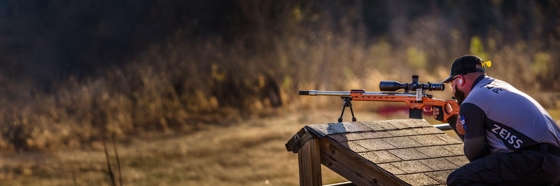 A person wearing a ZEISS-branded jacket is aiming a precision rifle equipped with a ZEISS scope from a wooden shooting platform in an outdoor setting.