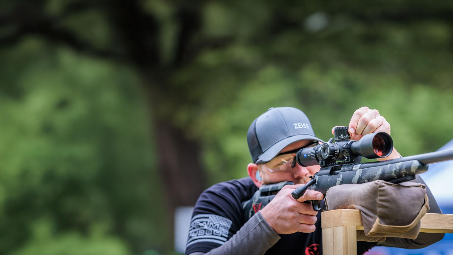 A person wearing a ZEISS cap adjusts the turret of a ZEISS riflescope mounted on a camouflage-patterned rifle.