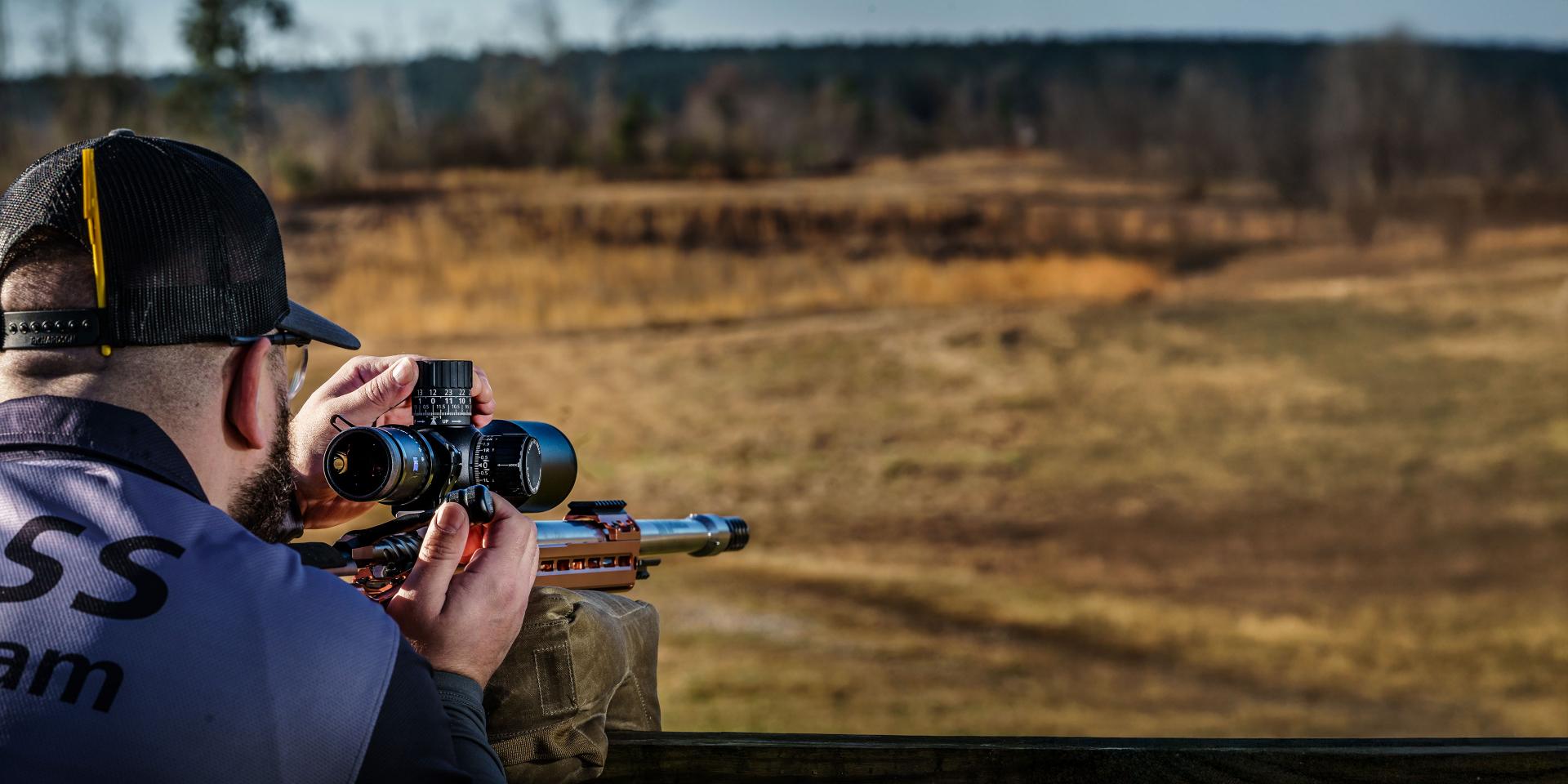 A man wearing a ZEISS team vest is aiming a rifle equipped with a ZEISS scope at a distant target in an open field.