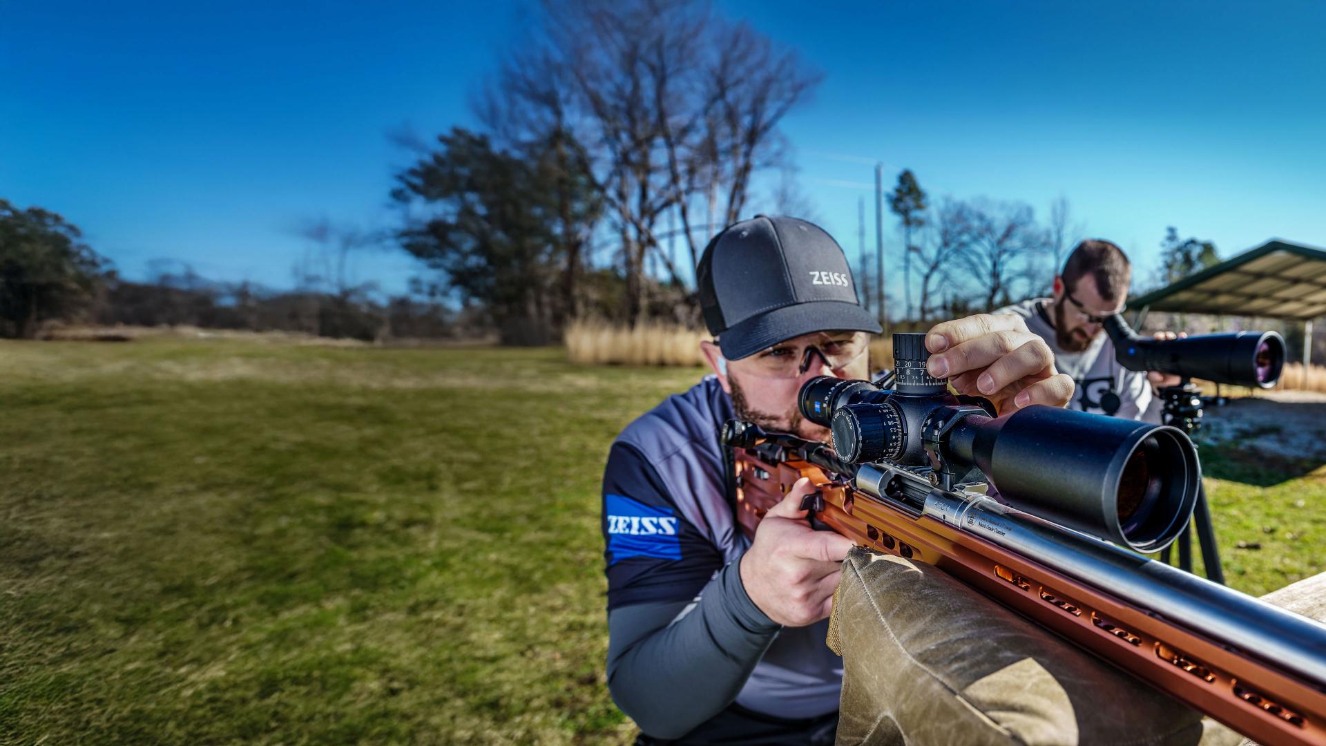 A marksman wearing ZEISS-branded gear aims through a ZEISS riflescope at an outdoor shooting range, with another person observing through a spotting scope in the background.