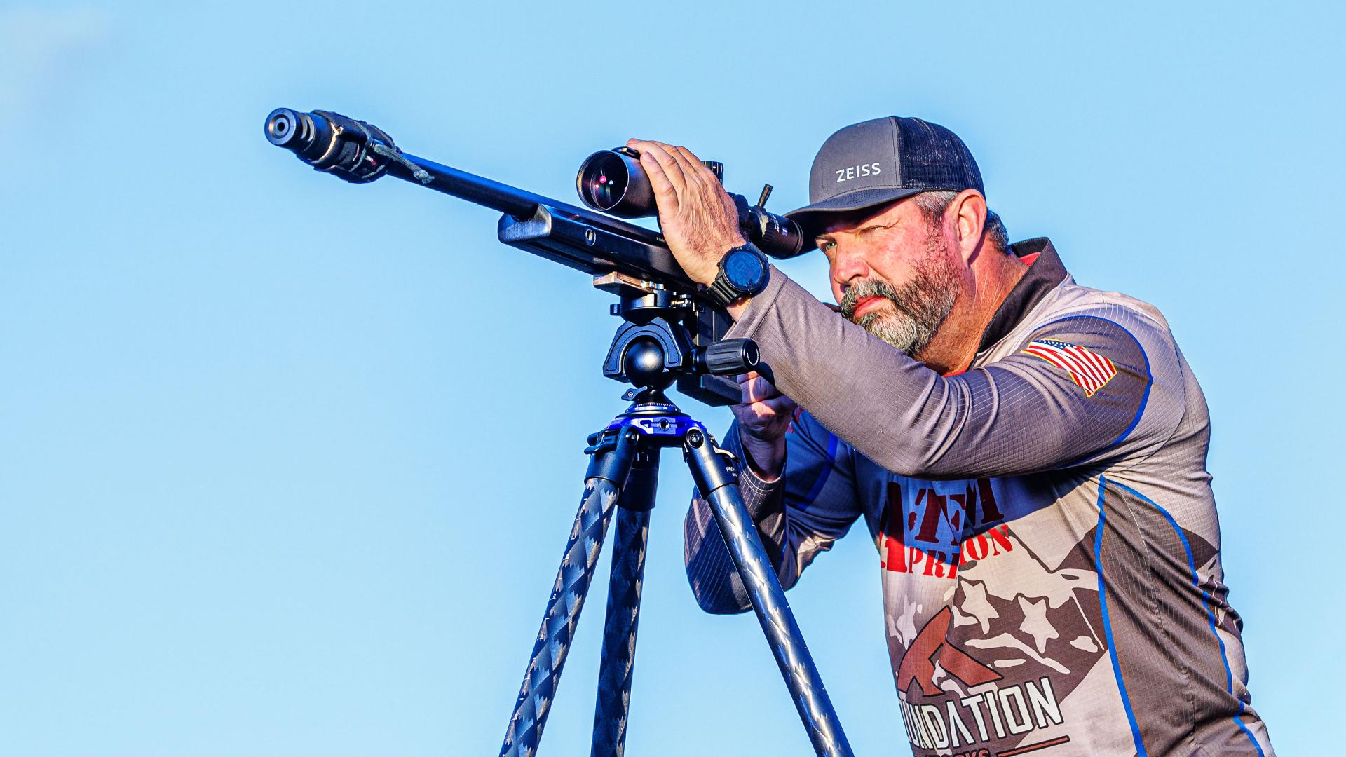 A man wearing ZEISS-branded gear aims a rifle equipped with a ZEISS scope mounted on a tripod against a clear blue sky.