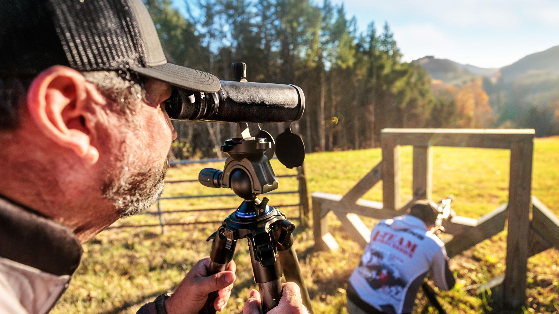 Man using a ZEISS spotting scope mounted on a tripod to observe a shooter aiming through a wooden structure in an outdoor field.