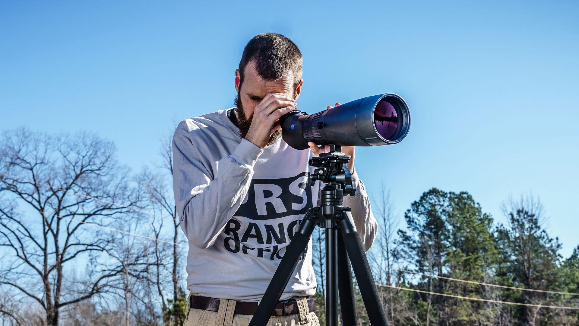A man is looking through a ZEISS spotting scope mounted on a tripod in an outdoor setting.