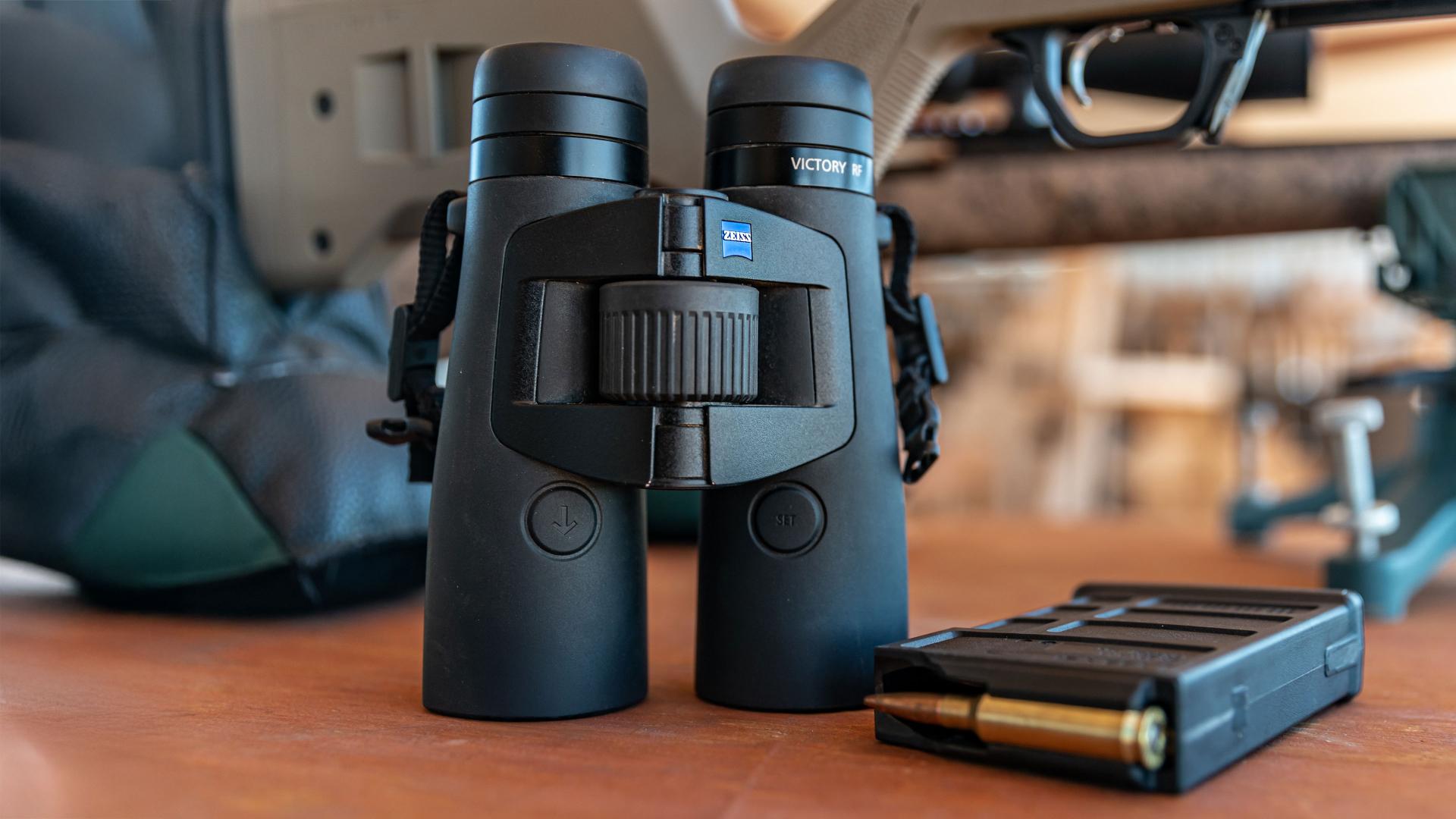 A pair of ZEISS binoculars placed upright on a wooden surface next to a loaded rifle magazine and part of a rifle.