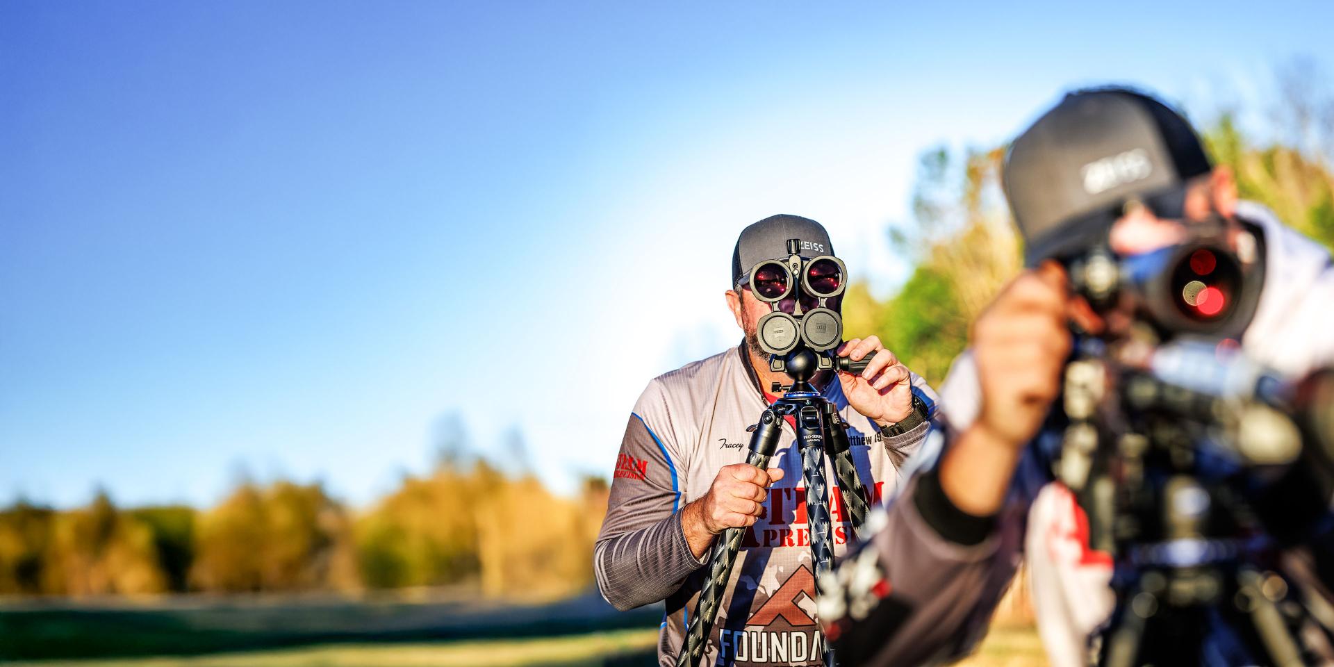Two individuals outdoors using ZEISS high-performance optics mounted on tripods, focused on distant targets in a natural landscape.