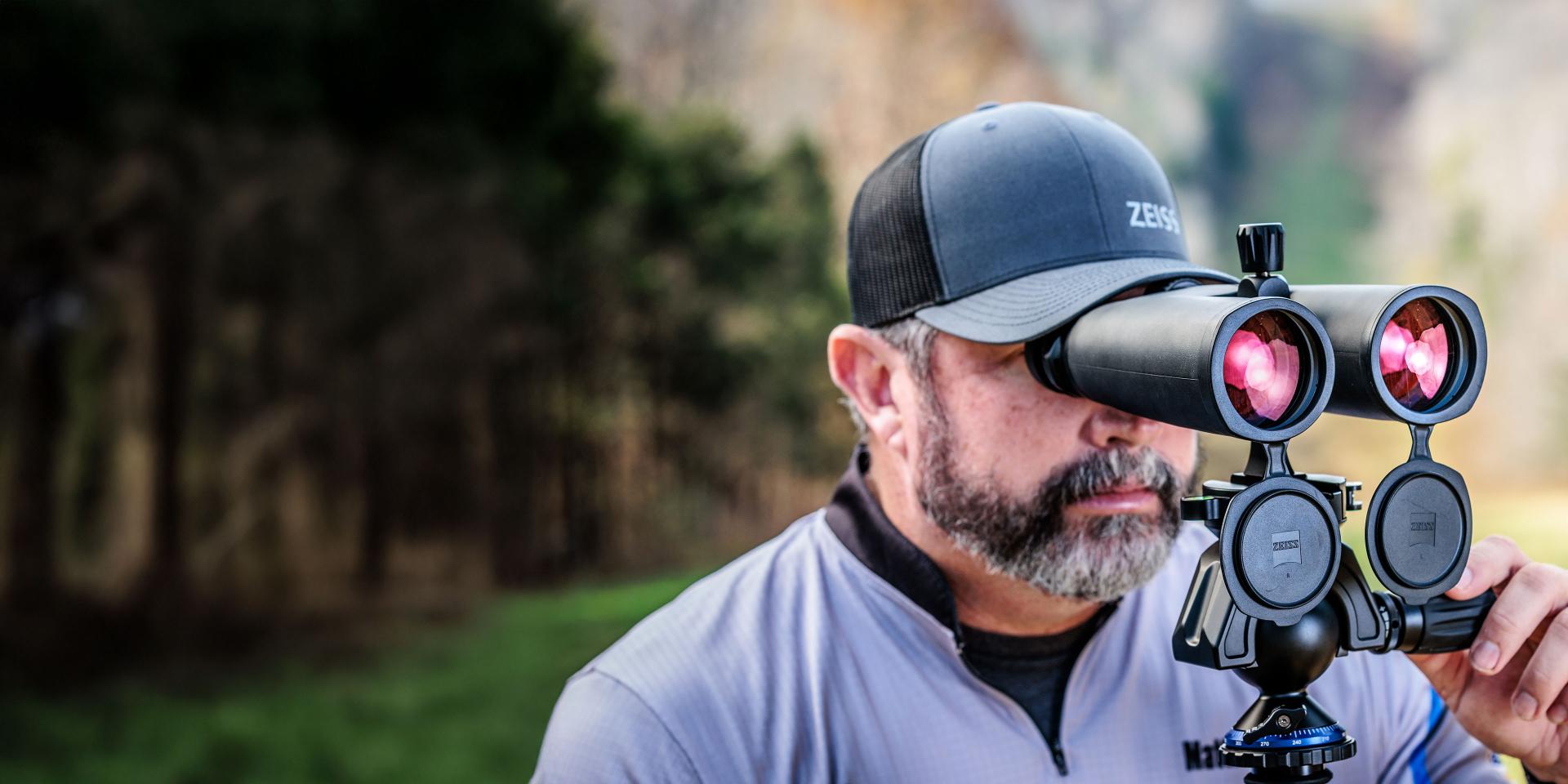 A man wearing a ZEISS cap uses a mounted pair of ZEISS binoculars with lens caps flipped open in an outdoor setting.