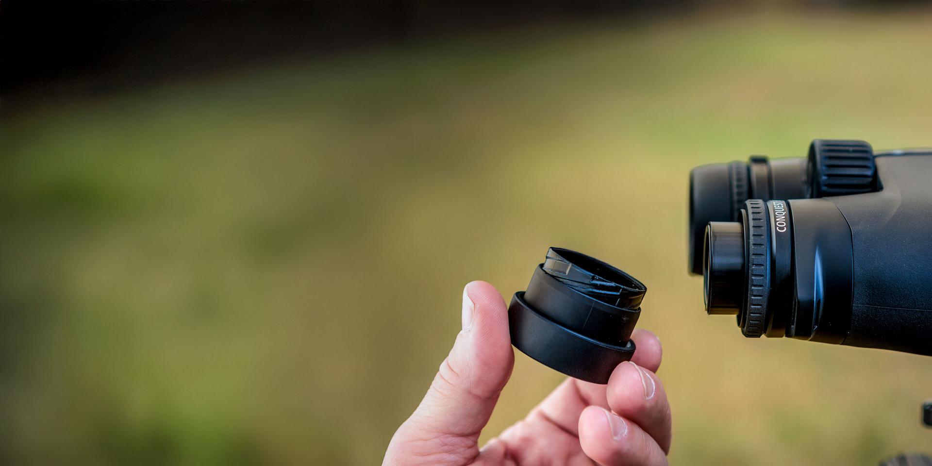 A person holds a removable eyepiece next to a pair of ZEISS binoculars against a blurred outdoor background.