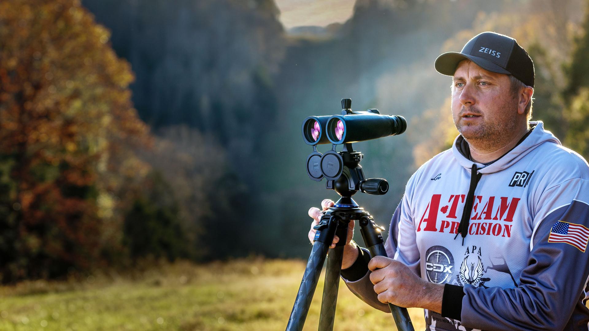 A man wearing a ZEISS cap is using a ZEISS rangefinder mounted on a tripod in a scenic outdoor setting with autumn trees.