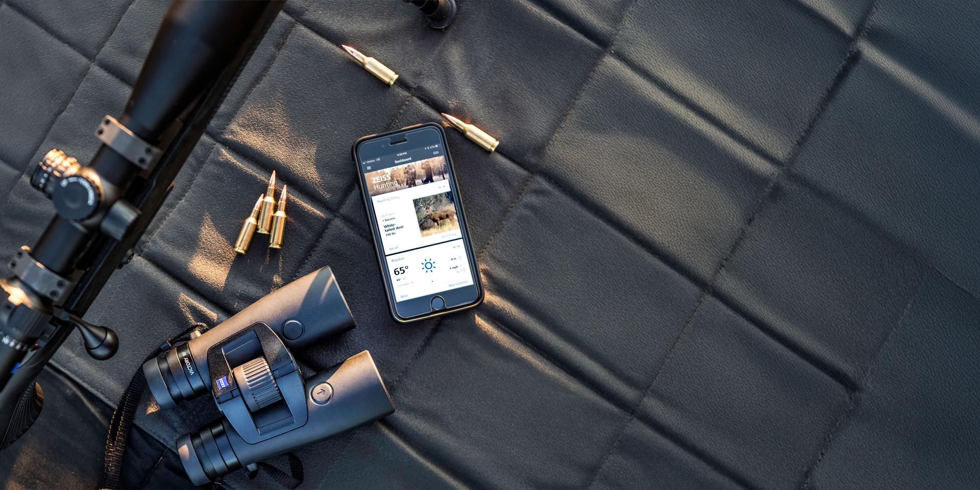 A ZEISS VICTORY RF binocular and a smartphone displaying the connected ZEISS app are placed on a tree stump in an outdoor forest setting.