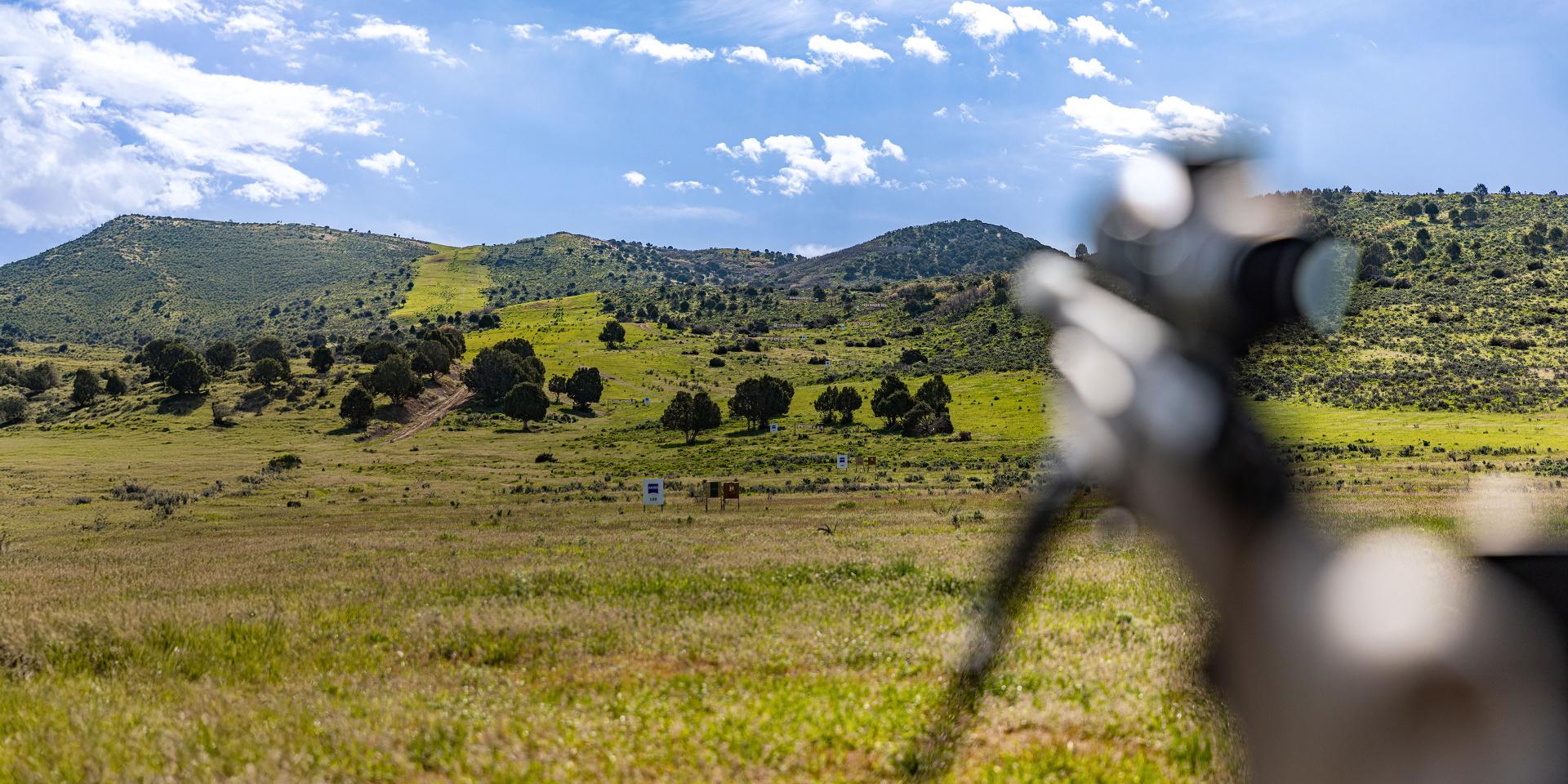 A blurred ZEISS riflescope in the foreground points toward a clear, sunlit landscape of grassy hills and scattered trees under a blue sky.