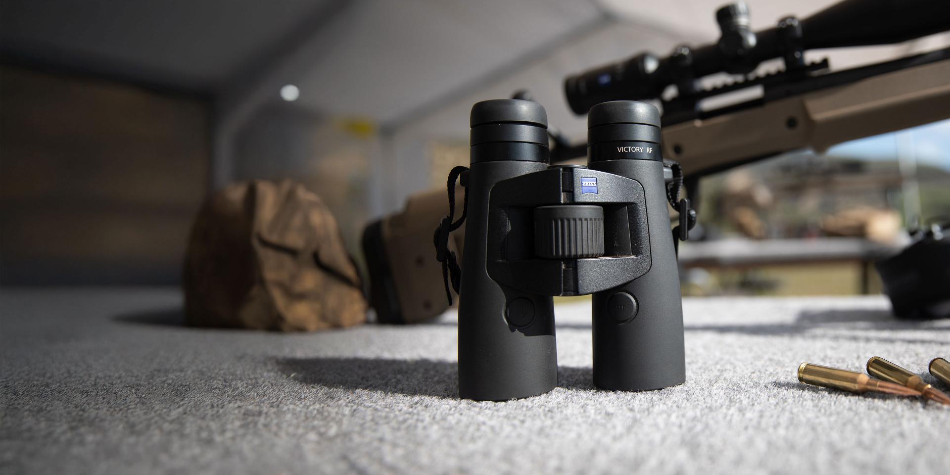 A precision rifle is displayed on a wooden surface alongside ZEISS binoculars, rifle cartridges, a cartridge box, and shooting accessories.