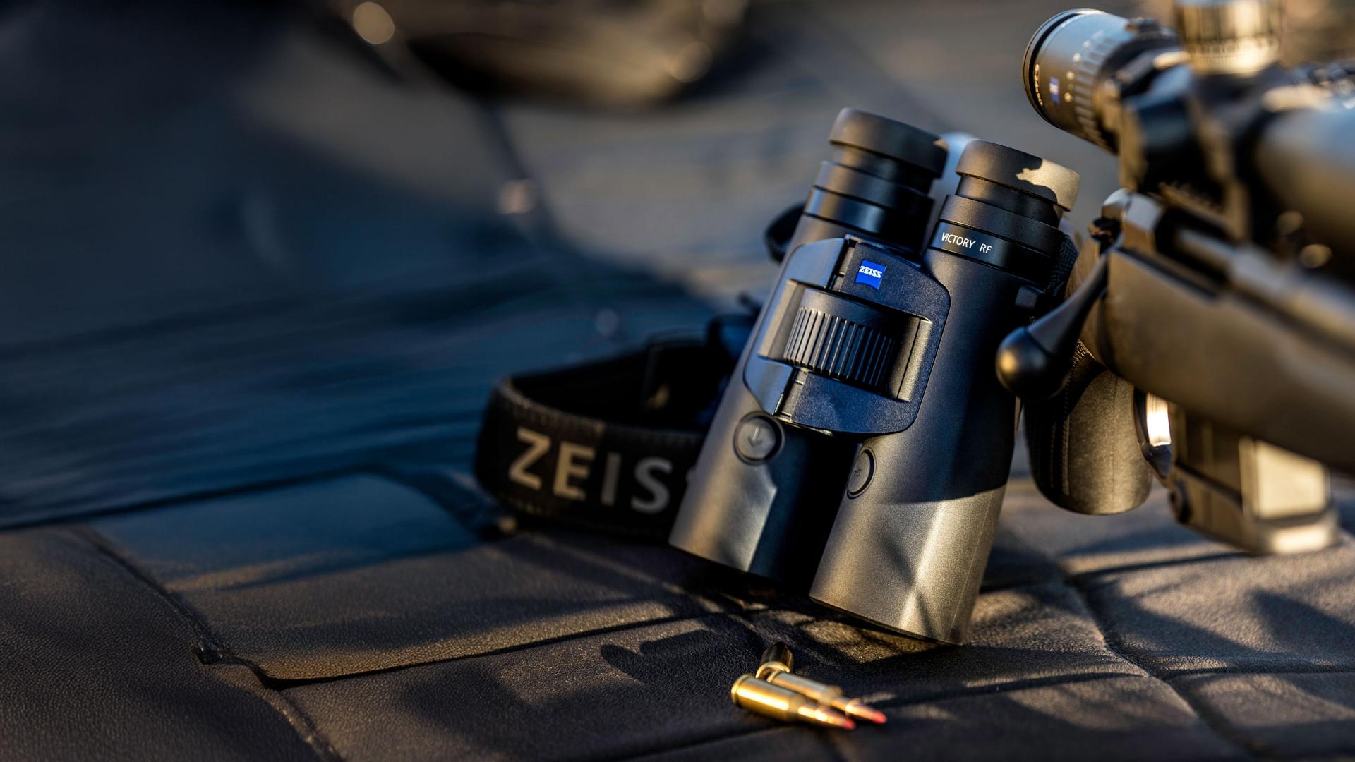 Black ZEISS binoculars with a ZEISS-branded strap are placed next to a rifle and two bullets on a padded surface in natural light.